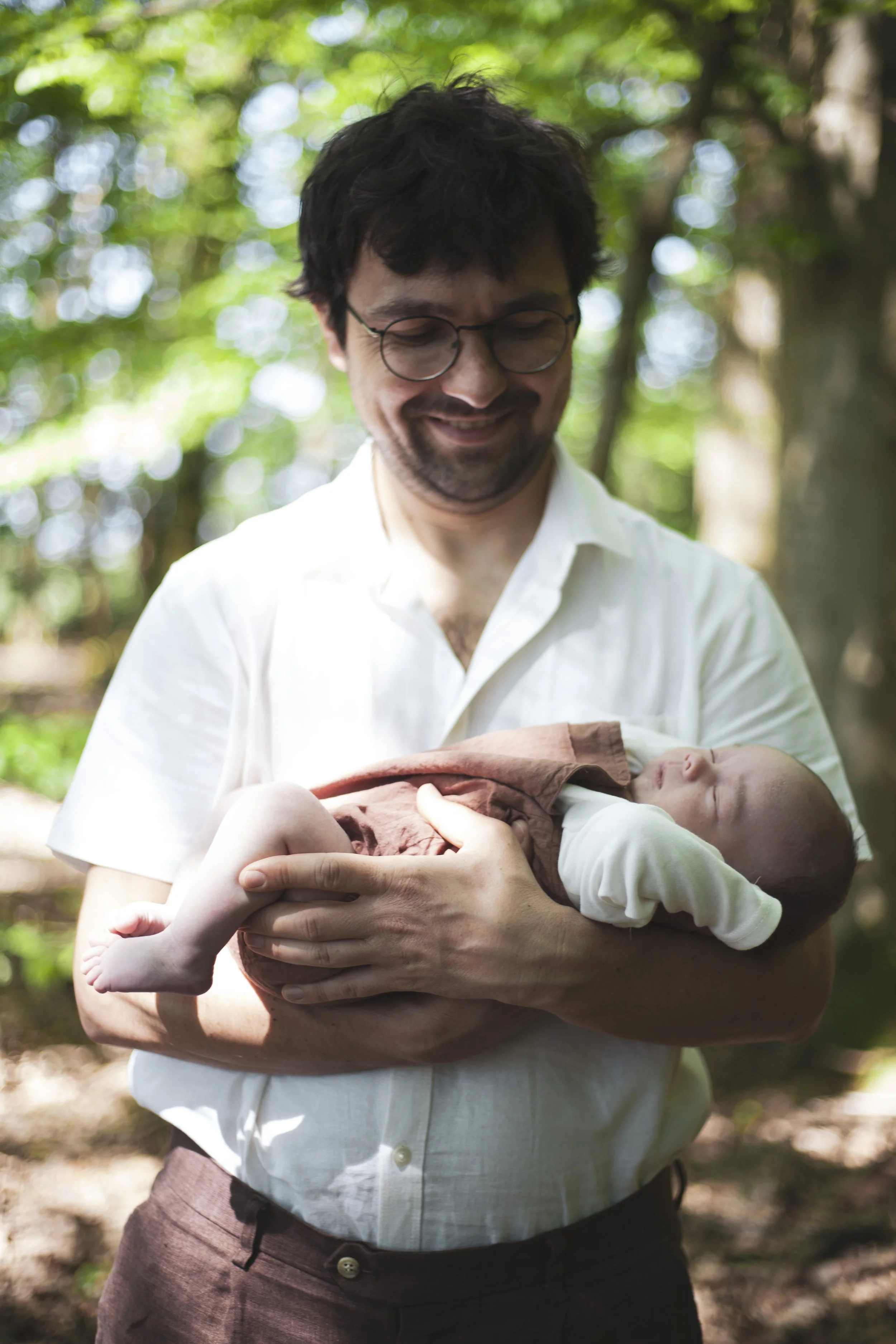 Ein Mann mit Brille hält ein schlafendes Baby im Arm in einem Wald, umgeben von grünen Bäumen. Newborn Photoshoot, Babyportrait, Fotografie. 