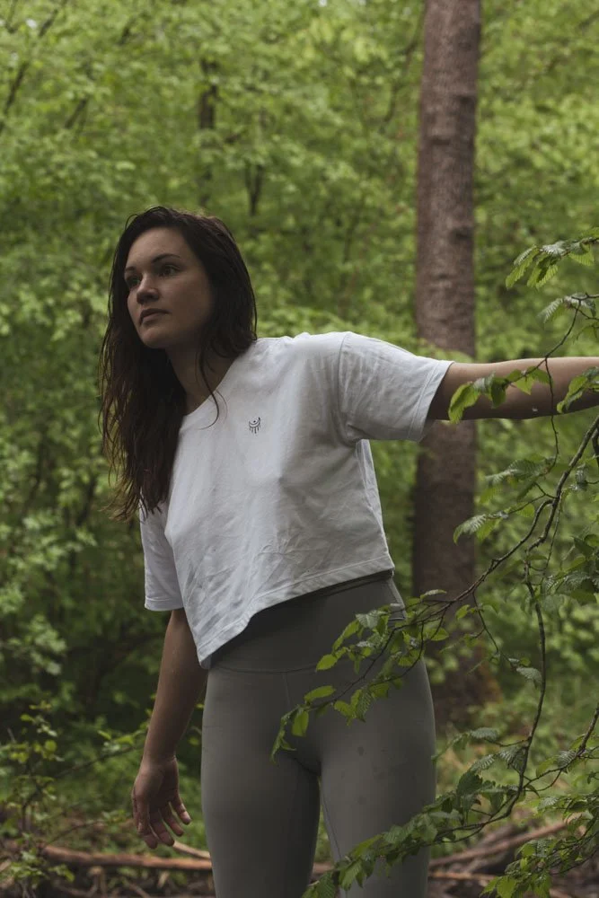 Eine junge Frau steht in einem grünen Wald und lehnt sich an einen Baum, trägt ein weißes T-Shirt und beige Leggings.