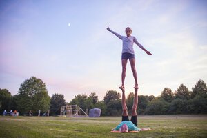Eine Person hält eine andere Person in der Luft auf einer Wiese, im Hintergrund Bäume und ein blauer Himmel mit wenigen Wolken.