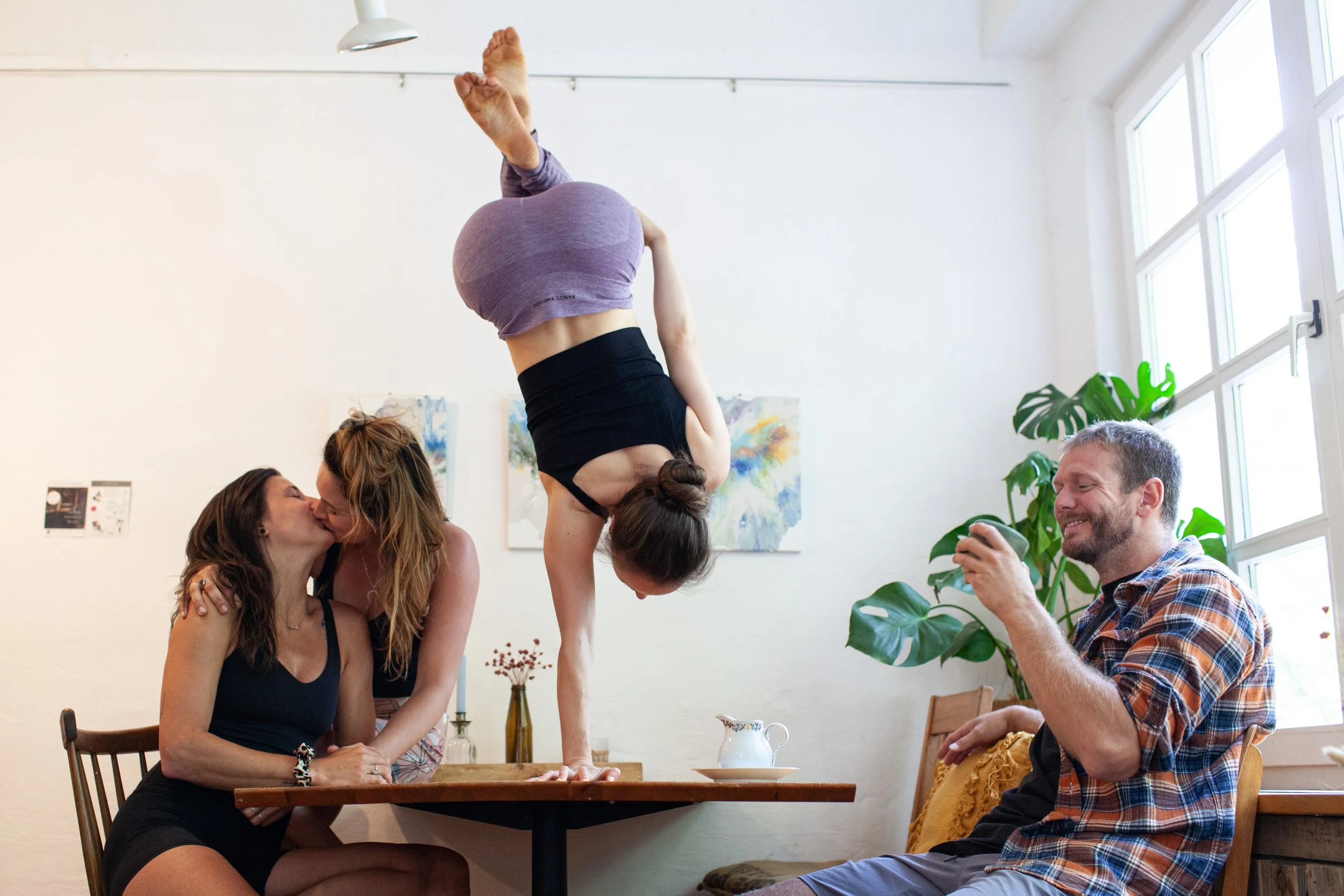 Mehrere Menschen im Wohnzimmer, eine Frau balanciert auf einem Tisch, während andere sitzen und fotografieren oder küssen, bei Tageslicht durch große Fenster, schöne Pflanzen im Hintergrund.