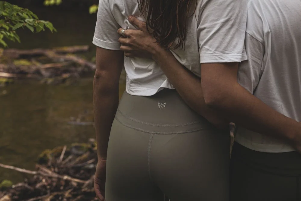 Nahaufnahme von zwei Menschen, die sich umarmen, beim Stehen am Fluss in der Natur, eine Person trägt graue leggings mit Logo am Rücken und der andere Person trägt ein weißes T-Shirt.