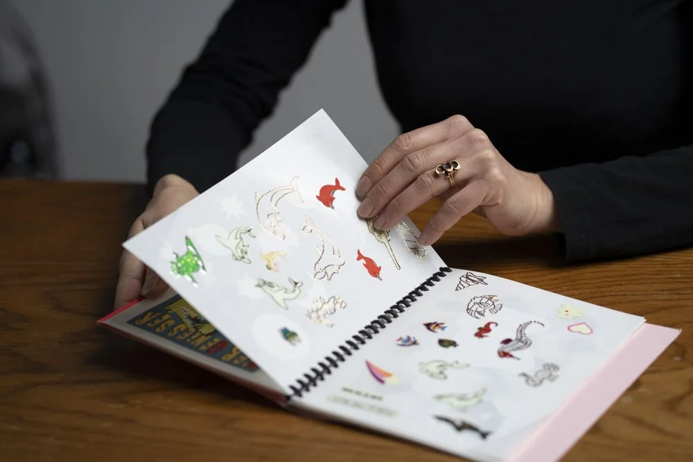 Person blättert in einem Stickeralbum mit Dinosaurieraufklebern auf einem Holztisch.