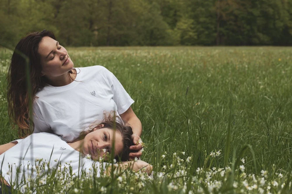 Zwei Frauen liegen in einer Wiese mit weißen Blumen, umgeben von Bäumen im Hintergrund, und genießen die Natur. Lifestyle, Fashionfotografie, Köln, Deutschland.