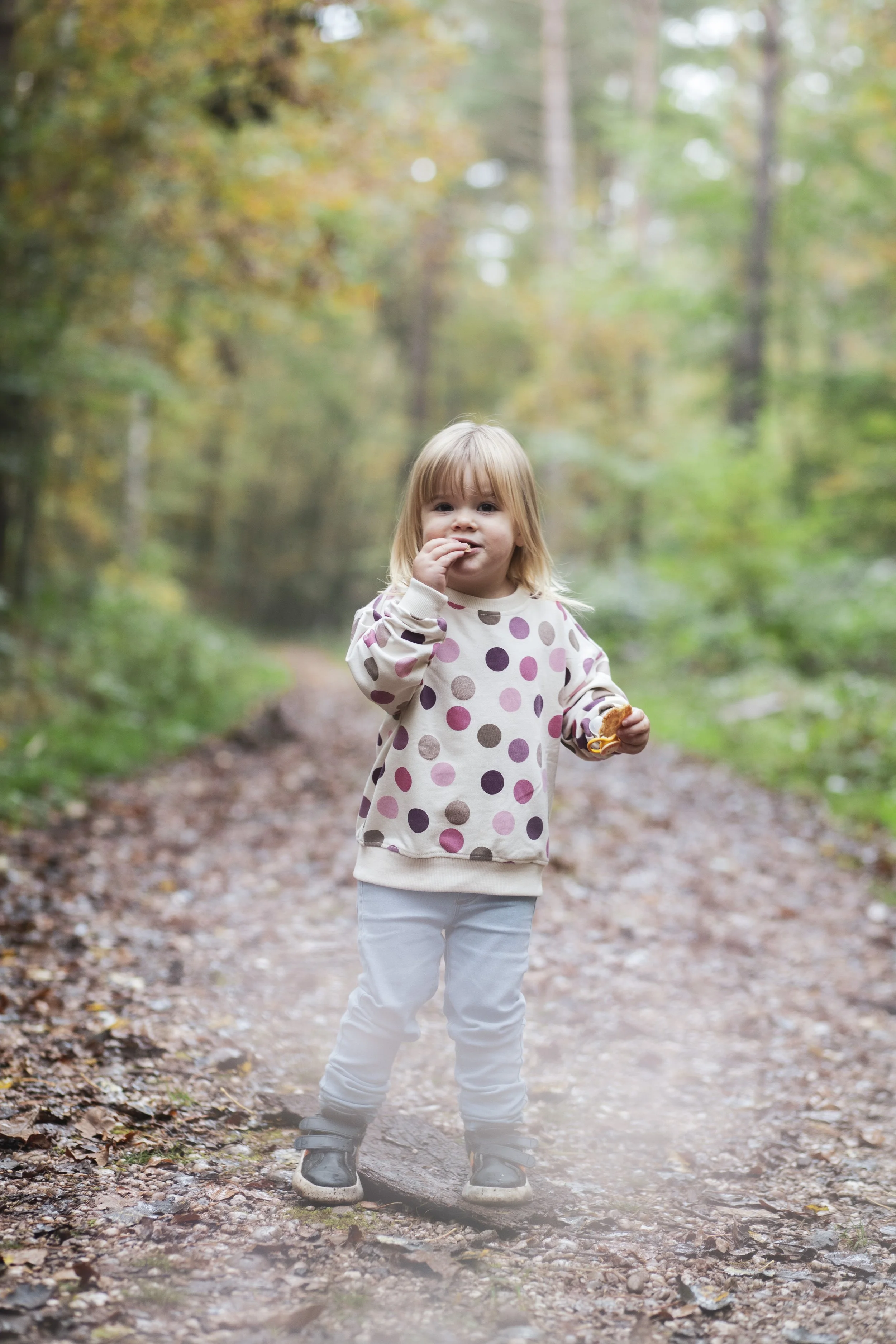 Ein kleines Mädchen mit blonden Haaren trägt einen Pullover mit bunten Punkten, blaue Hosen und Schuhe, es steht auf einem Weg im Wald, umgeben von herbstlichen Bäumen und Laub. Kinderportrait, Naturfotografie, Familienportrait.