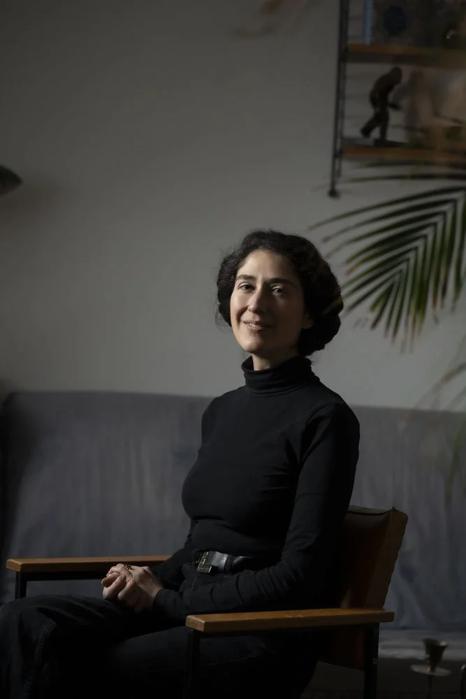 Frau mit dunklem, lockigem Haar sitzt in einem grauen Raum, trägt schwarzen Rollkragenpullover und lächelt leicht. Portrait, Saarbrücken, Jessica Bruni. 