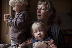 Eine Frau hält ein kleines Kind im Arm, während ein älteres Kind neben ihnen steht und auf etwas in den Händen schaut.