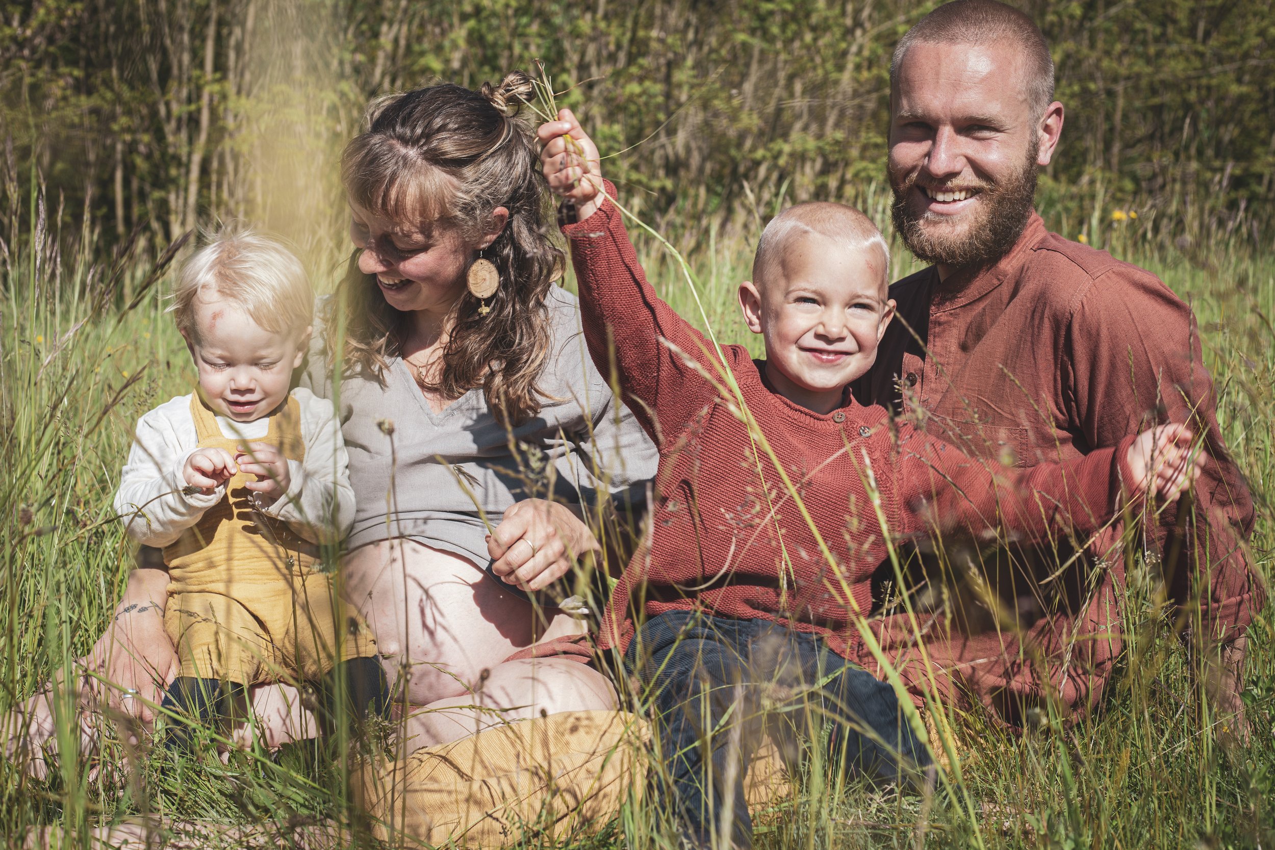 Eine glückliche Familie mit zwei Kindern sitzt im hohen Gras in einem Wald, die Mutter und der Vater lachen, während die Kinder spielen. Familienportrait, Natur, Kinderportraits. 