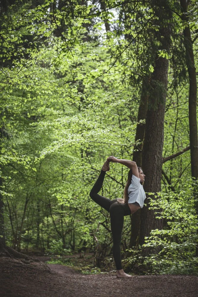 Frau führt Yoga im Wald aus, umgeben von grünen Bäumen, mit Baum im Hintergrund.