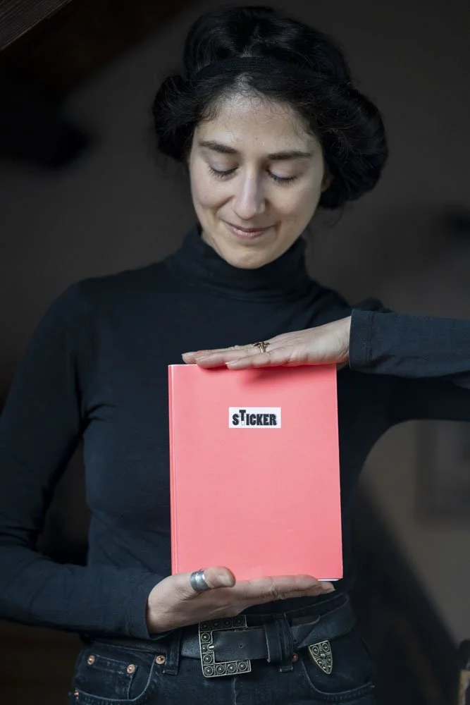 Eine junge Frau mit dunklem, gewelltem Haar trägt einen schwarzen Rollkragenpullover und hält ein rosa Notizbuch mit einem schwarzen und weißen Aufkleber mit der Aufschrift 'STICKER'. Sie lächelt leicht und schaut auf das Notizbuch.