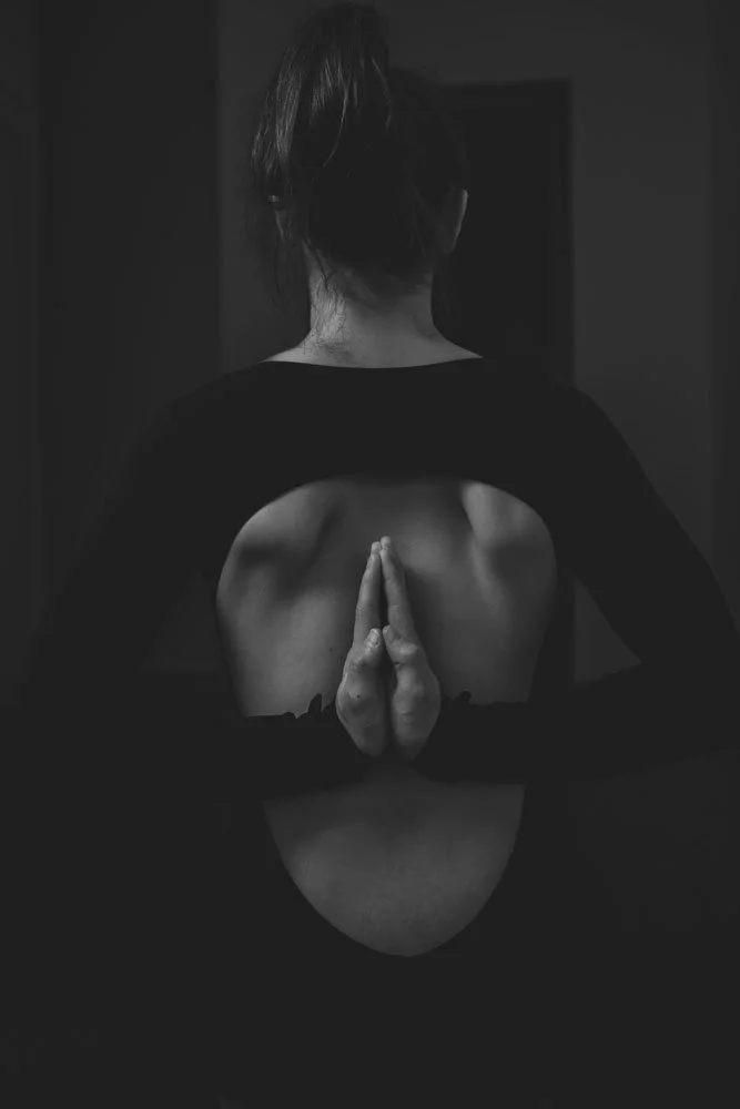 Schwarzes und weißes Foto einer Person, die eine Yoga-Position macht, mit einem Blick auf den Rücken, die Schultern und Hände in Gebetsposition hinter dem Rücken.