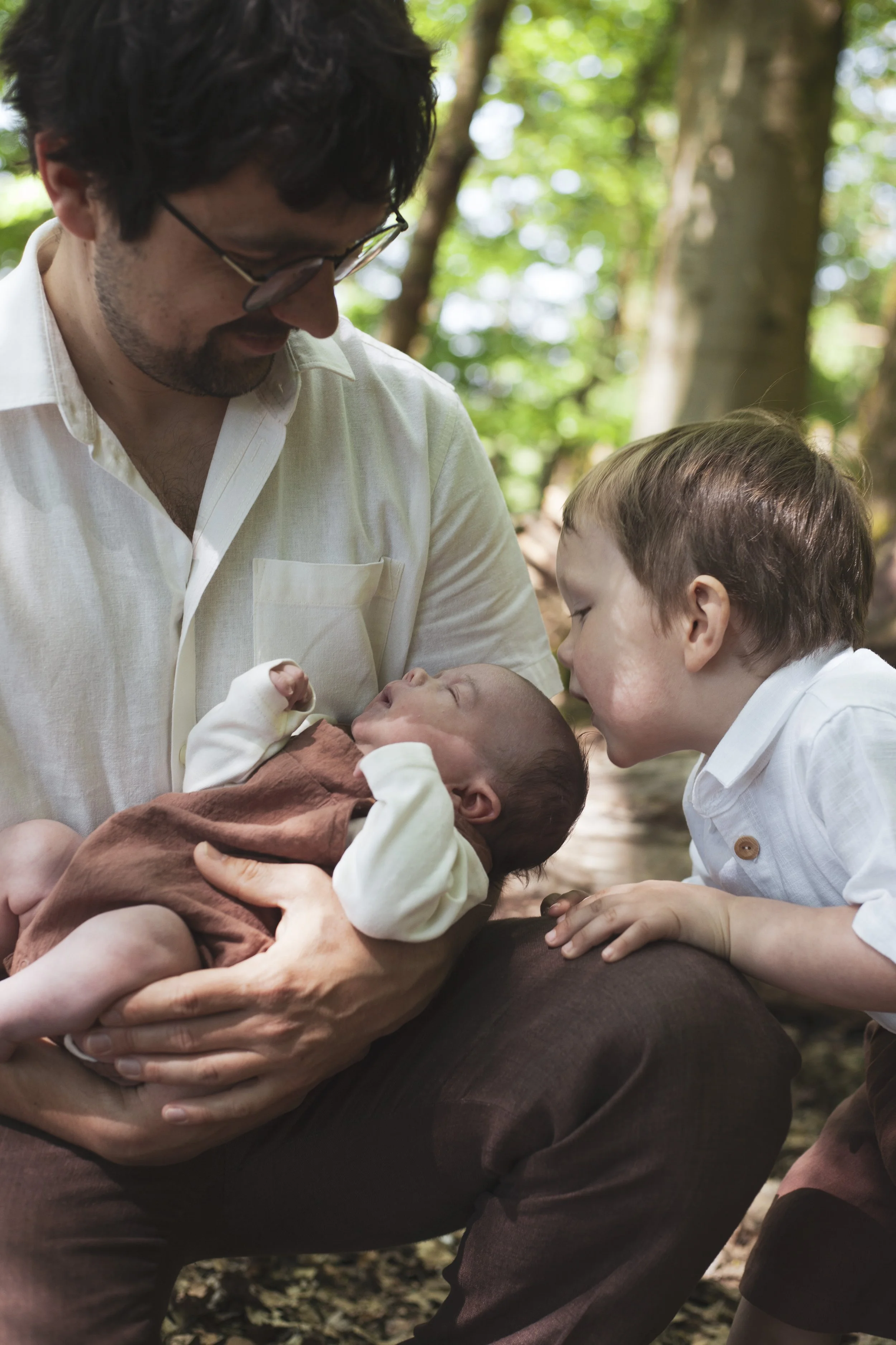 Ein Mann hält ein neugeborenes Baby im Arm, während ein älterer Junge neugierig schaut, in einem Wald mit Sonnenlicht und grünen Blättern im Hintergrund.