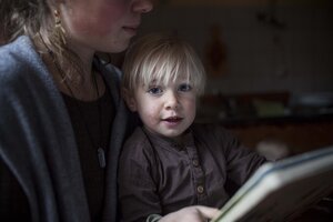 Ein kleines Kind sitzt auf dem Schoß einer Frau, die vermutlich seine Mutter ist, und schaut auf ein Tablet, in einem dunklen Raum.