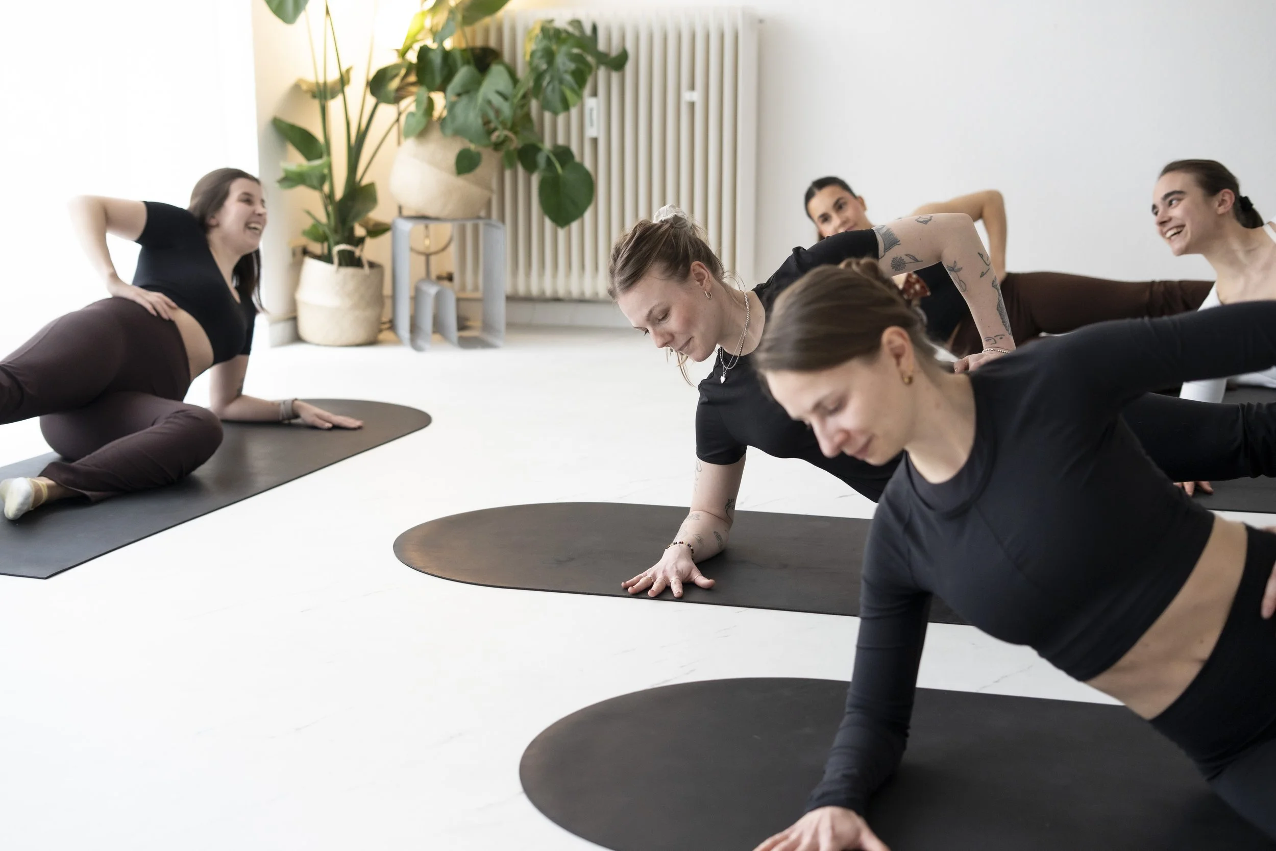 Menschen nehmen an einem Yoga-Kurs in einem hellen Raum teil, sitzen auf schwarzen Yogamatten und lachen während einer Übung.
