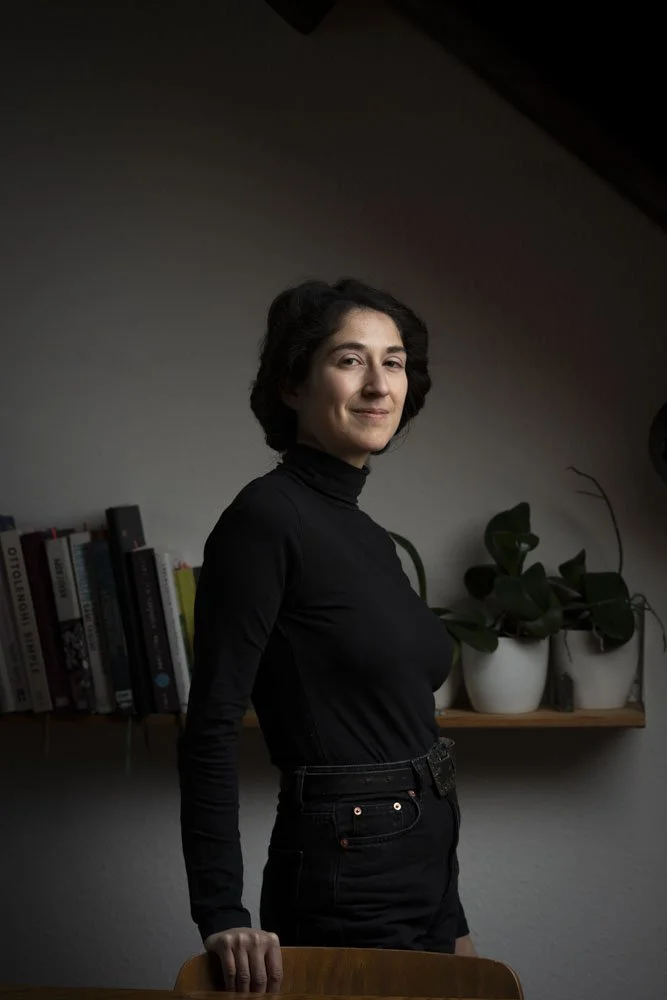Eine junge Frau mit dunklen, lockigen Haaren, trägt ein schwarzes Langarmshirt und schwarze Hose, steht in einem Raum mit Bücherregal und Pflanzen im Hintergrund.