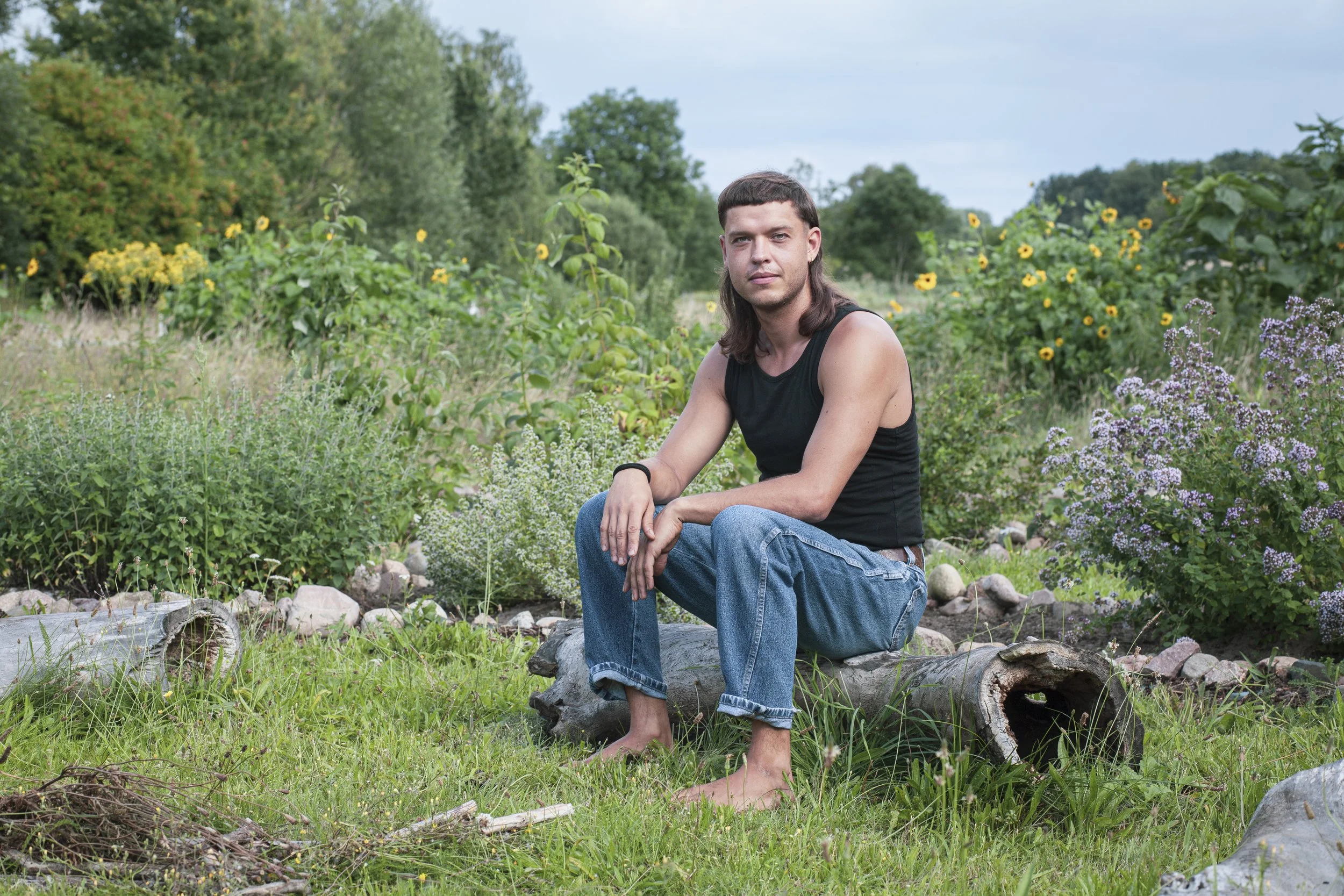 Ein junger Mann mit langen Haaren, schwarzen Tanktop und Jeans sitzt barfuß auf einem Baumstamm in einem Garten mit vielen Pflanzen und Blumen, im Hintergrund Bäume und ein bewölkter Himmel.