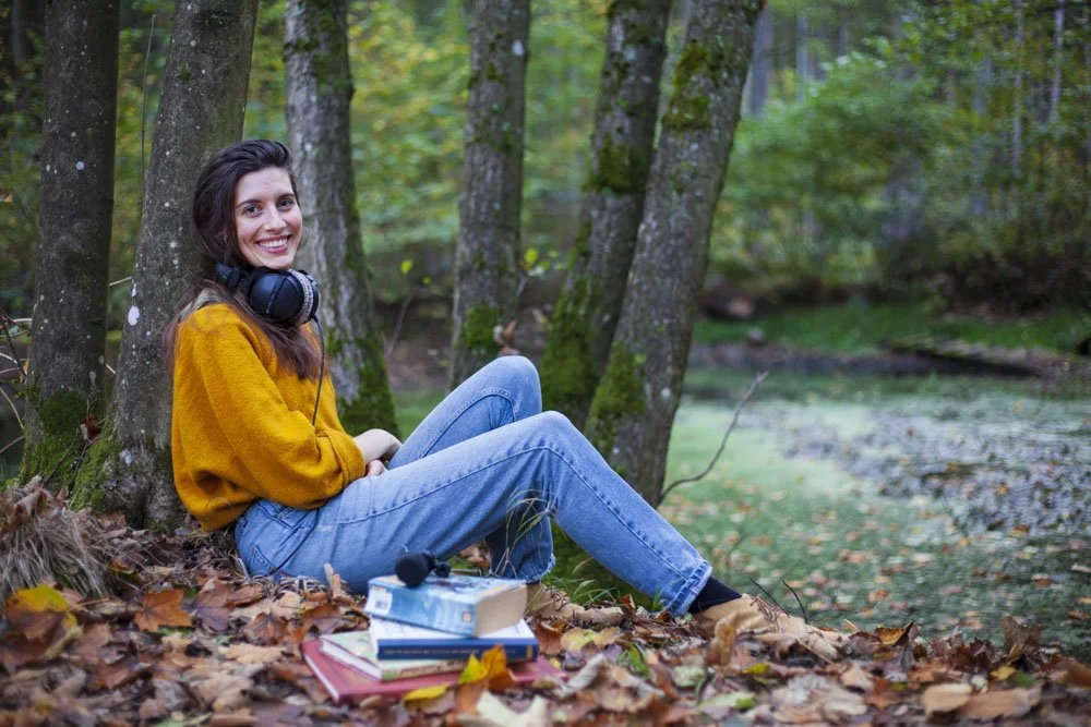 Eine lachende junge Frau im herbstlichen Wald sitzt auf Laub neben Bäumen, mit Kopfhörern um den Hals und Büchern vor ihr.