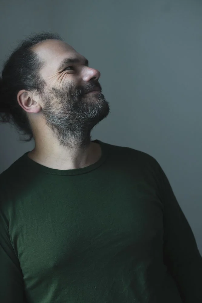 Ein lachender Mann mit dunklem Haar, Bart und Bartstoppeln trägt ein schwarzes T-Shirt und steht vor einem grauen Hintergrund.