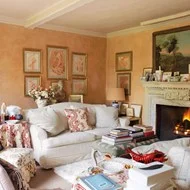 This living room designed by Nicky Haslam has all the hallmarks of coziness. From the pair of comfortable sofas facing the fireplace, abundance of cushions, fabric ottoman to either put your feet up or hold stacks of books, to the intricately patterned floor-length tablecloth. The coral hand-glazed wall finish and selection of wall art in gilt frames pulls all the elements of the room together. The silk lampshade complements the walls and will cast a warm glow when lit.
