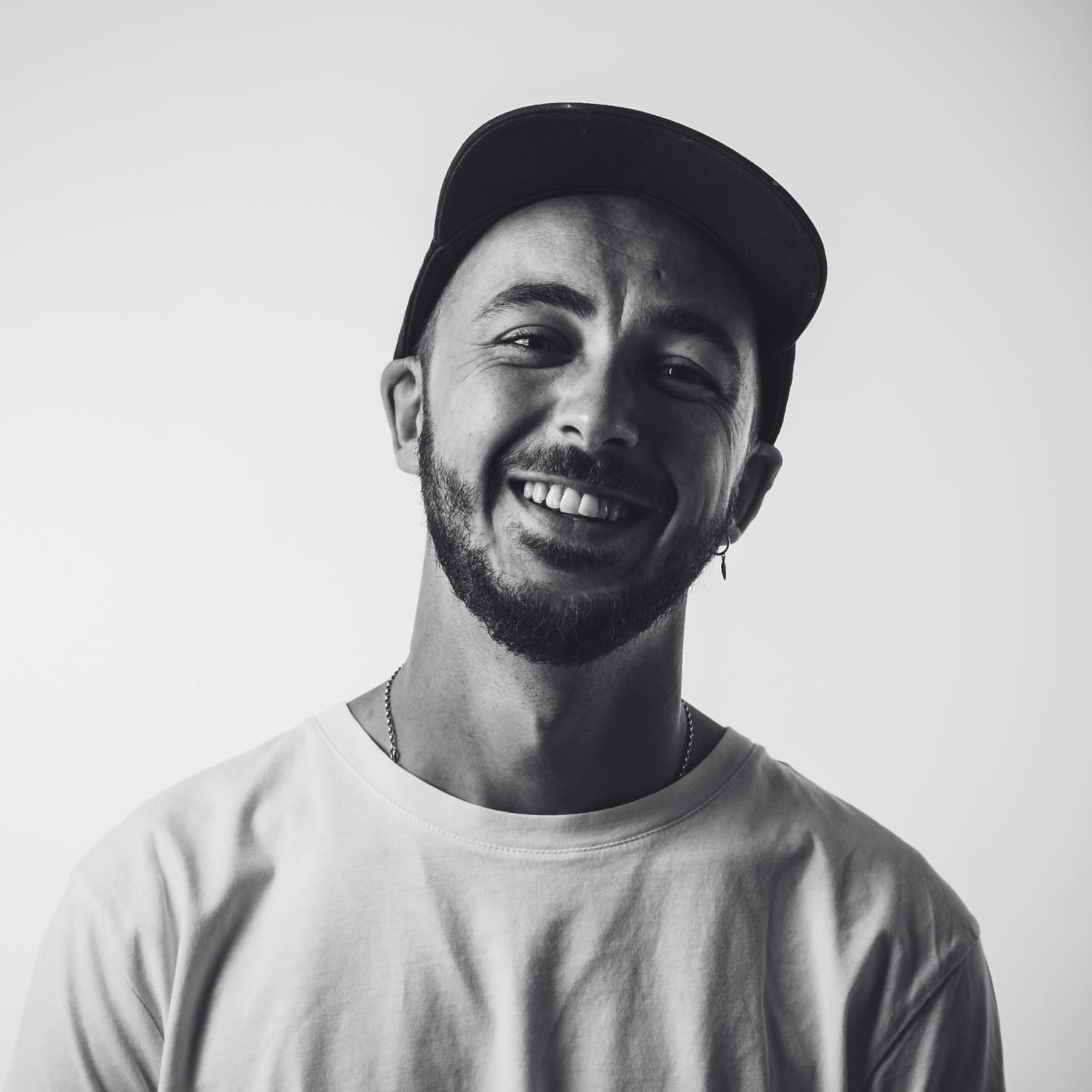 Black and white photo of a smiling man wearing a baseball cap, earrings, a necklace, and a light-colored T-shirt.