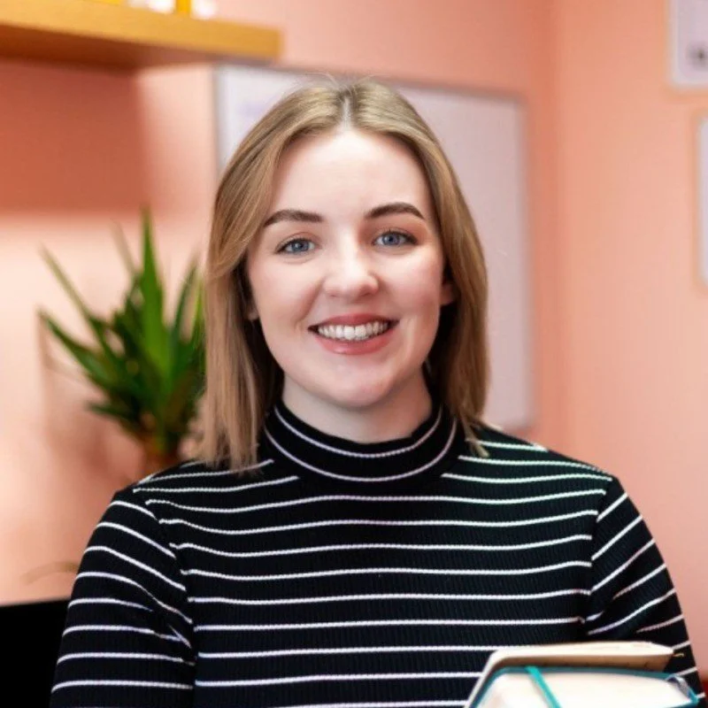 A smiling young woman with shoulder-length blonde hair in a black and white striped turtleneck, standing in an indoor setting with a peach-colored wall and a potted green plant in the background.
