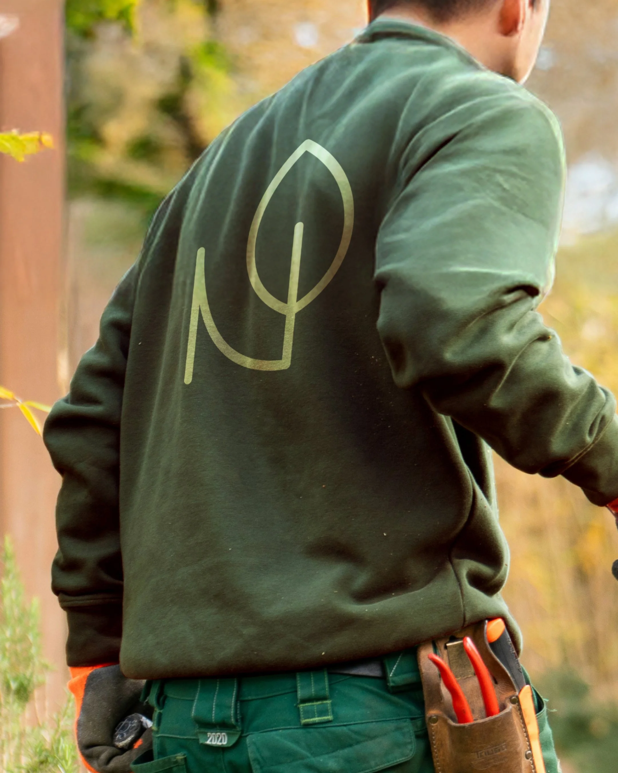 Person with brown hair wearing a green sweatshirt with a leaf logo on the back and green work pants, outdoors with blurred autumn foliage in the background.