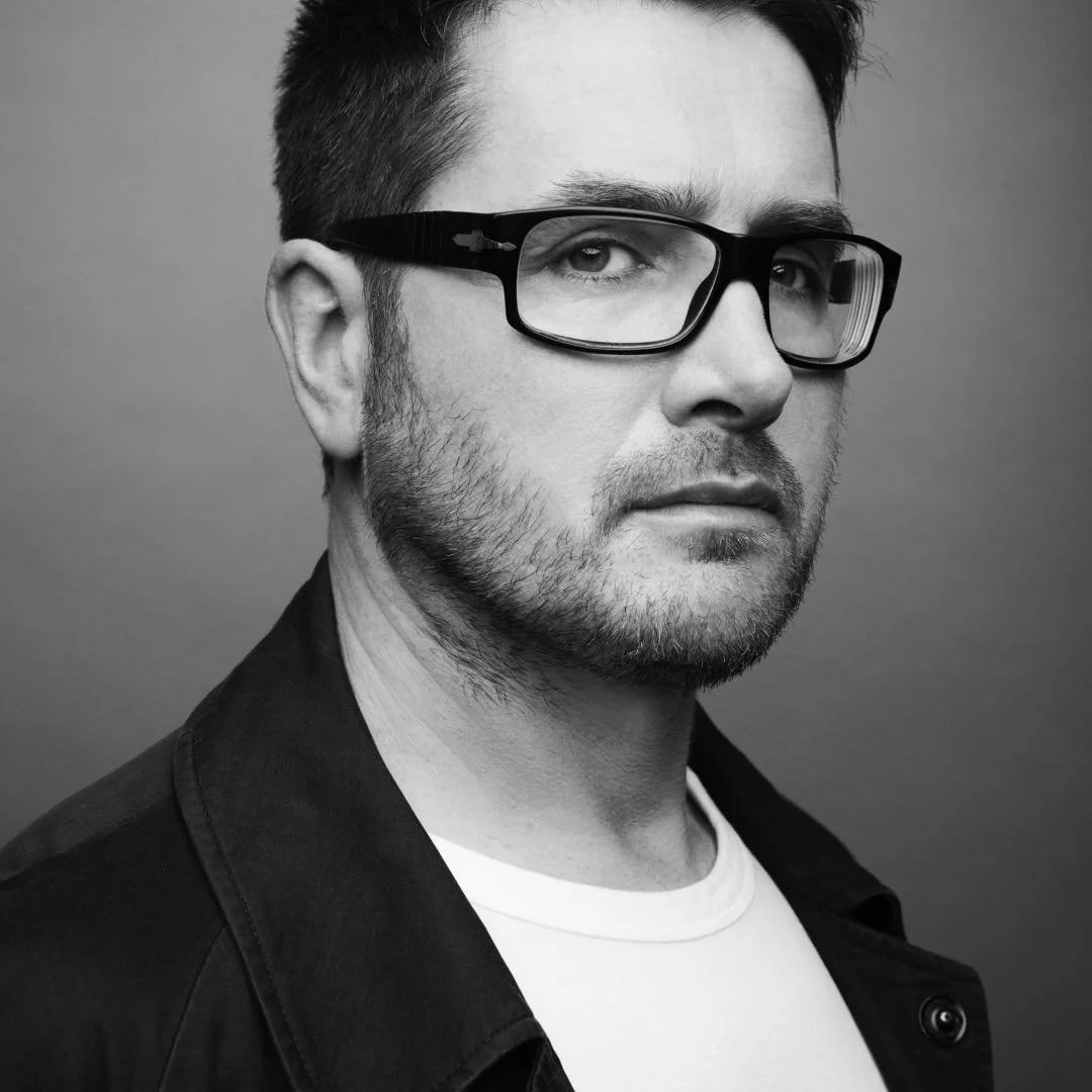 Black and white portrait of a man with light skin, short dark hair, glasses, slight beard, wearing a dark jacket and a light shirt, looking at the camera with a neutral expression.