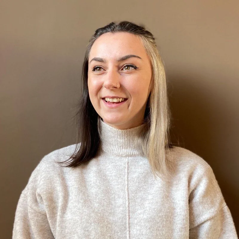 A young woman with shoulder-length hair, one side black and the other blonde, smiling and looking to the side, wearing a cream-colored turtleneck sweater against a plain beige wall.
