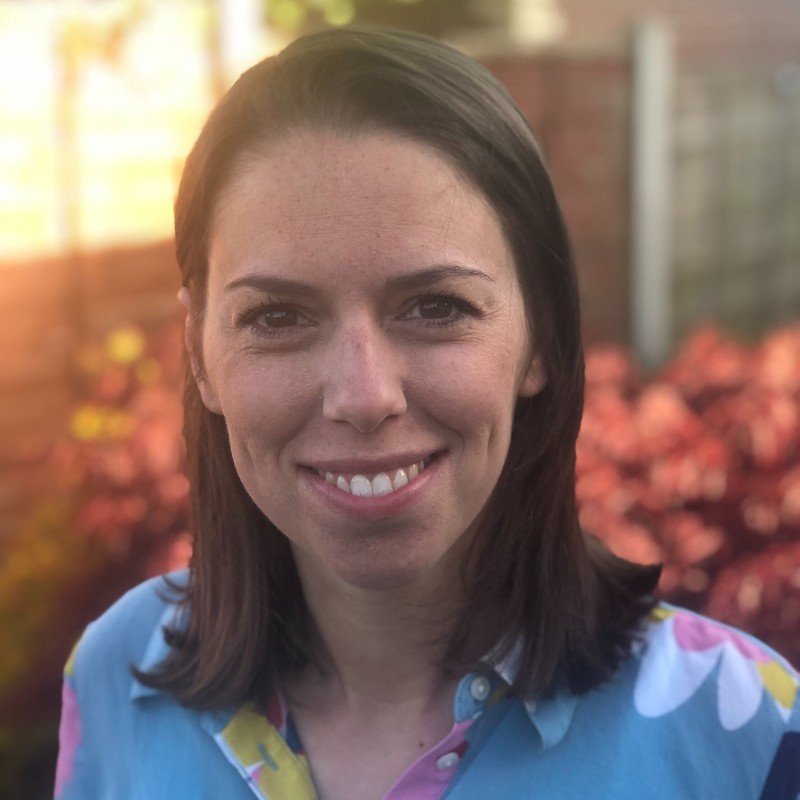 A woman with shoulder-length dark hair smiling outdoors with pink flowers and a wooden fence in the background.