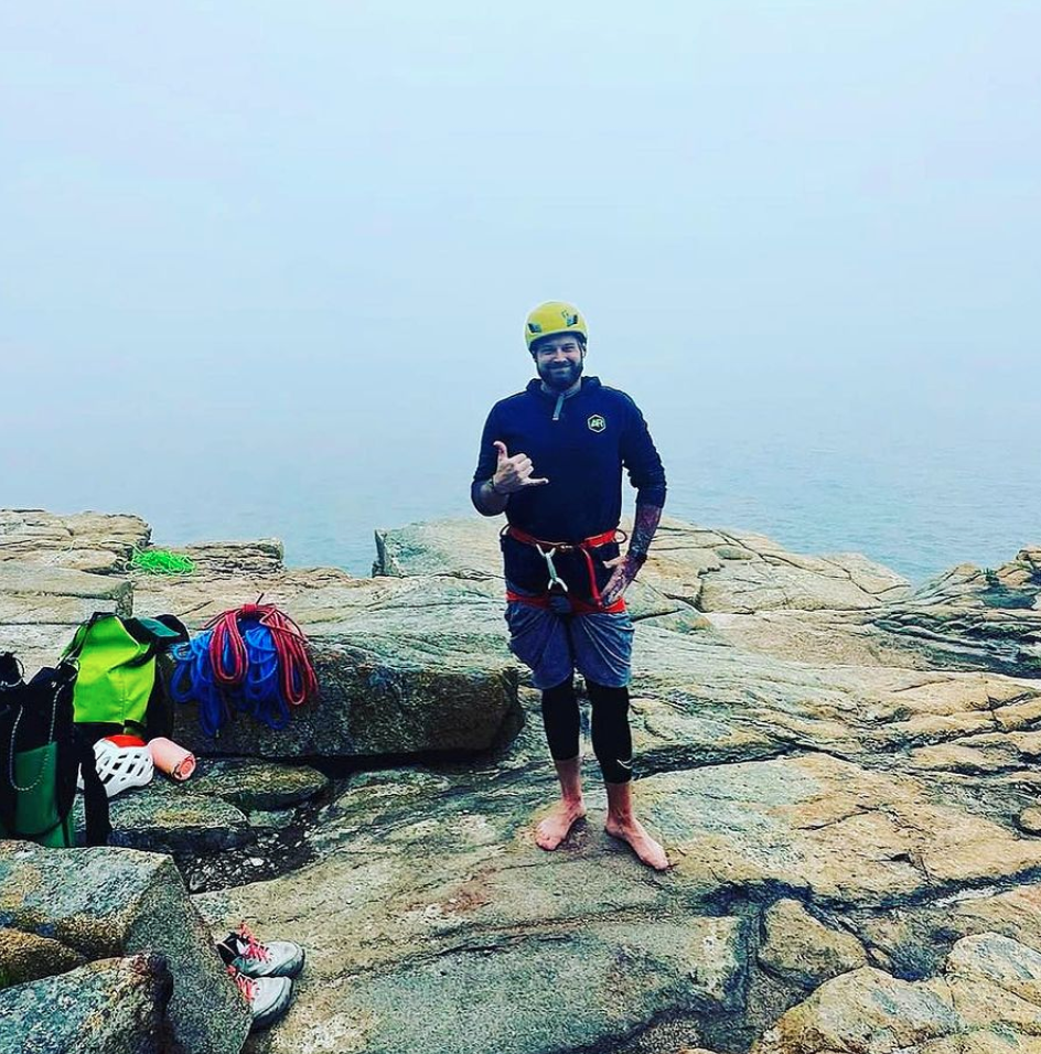 Josh Flaherty, LCSW, executive director of Adventure Recovery, stands barefoot on rocky terrain next to the ocean, smiling and making a shaka gesture. Climbing gear and shoes are placed on the rocks nearby.