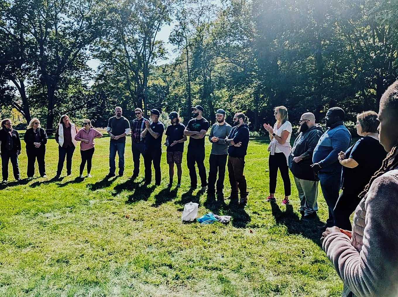 Group of people standing in a semi-circle outdoors on a sunny day, with trees in the background.