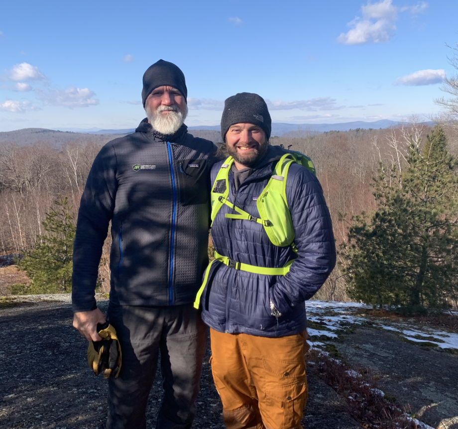 Tim Walsh, founder of Adventure Recovery, and Josh Flaherty, LCSW, in outdoor gear standing on a rocky hilltop with a view of a forest and mountains in the background