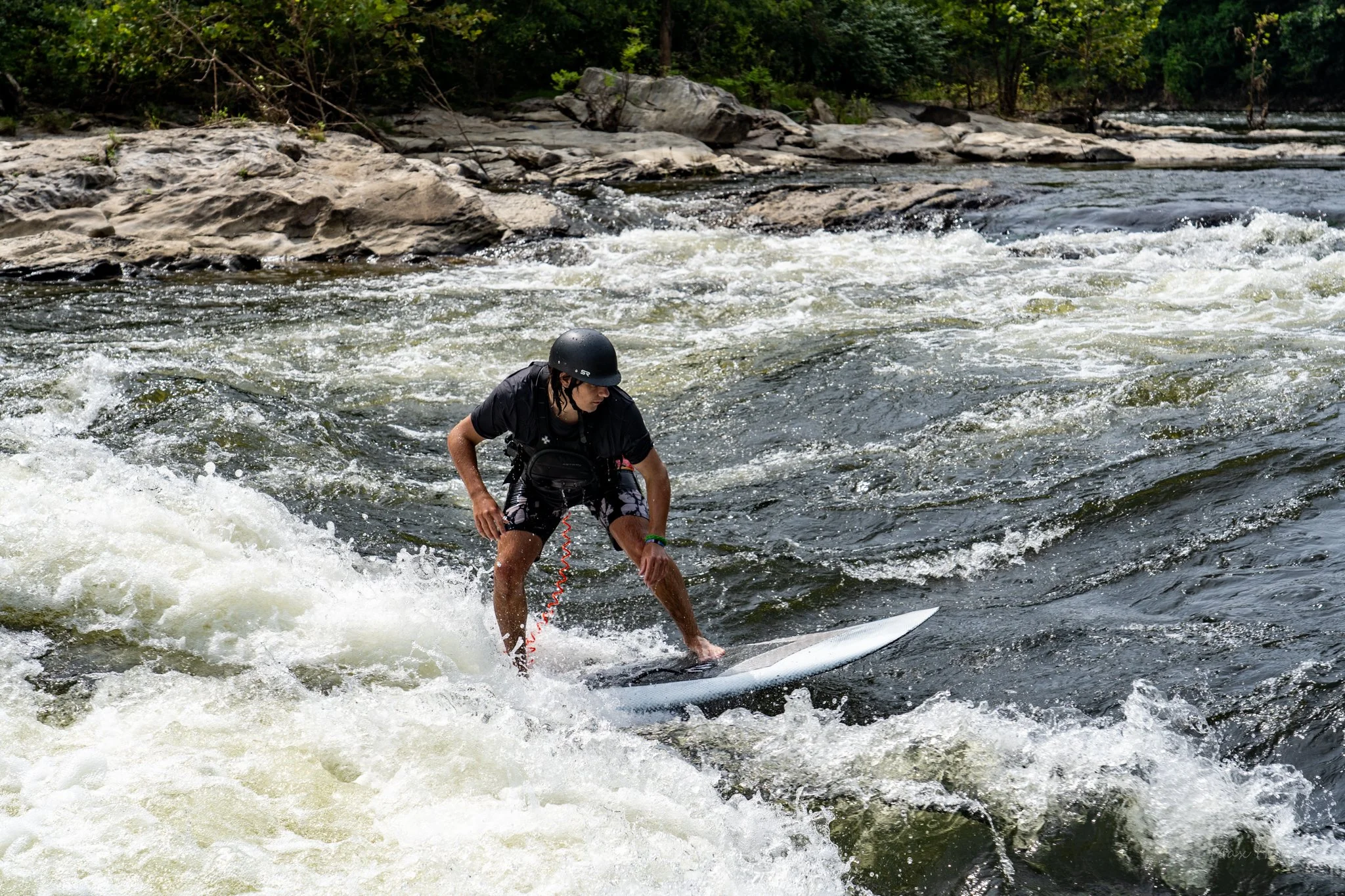 A person wearing a black helmet and black clothing is surfing on a river with white water rapids, surrounded by rocks and green trees.