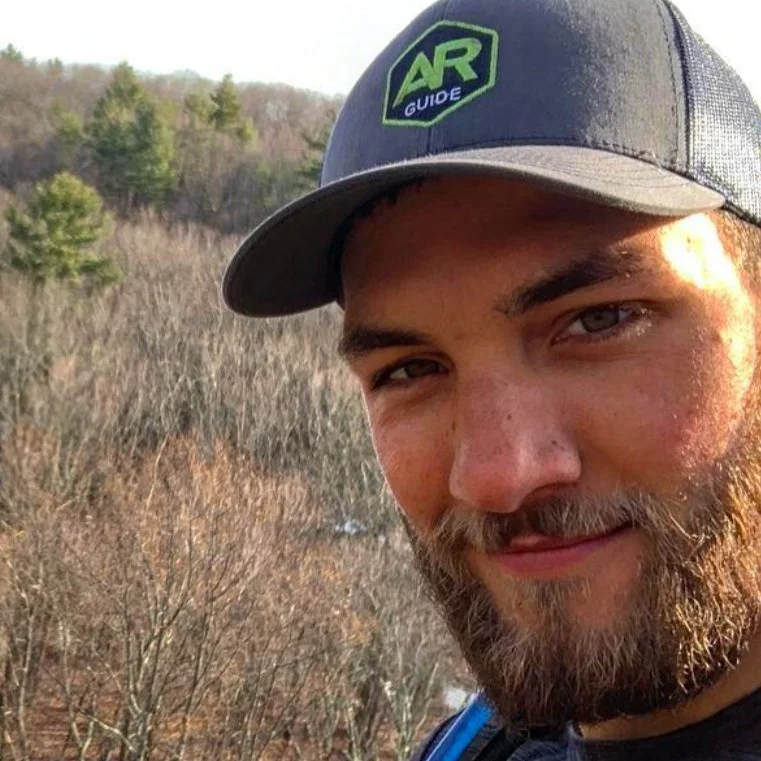 Close-up of a young man with a beard wearing a black cap that has a green and blue logo reading "AR Guide." He is outdoors with bare trees and some evergreen trees in the background.