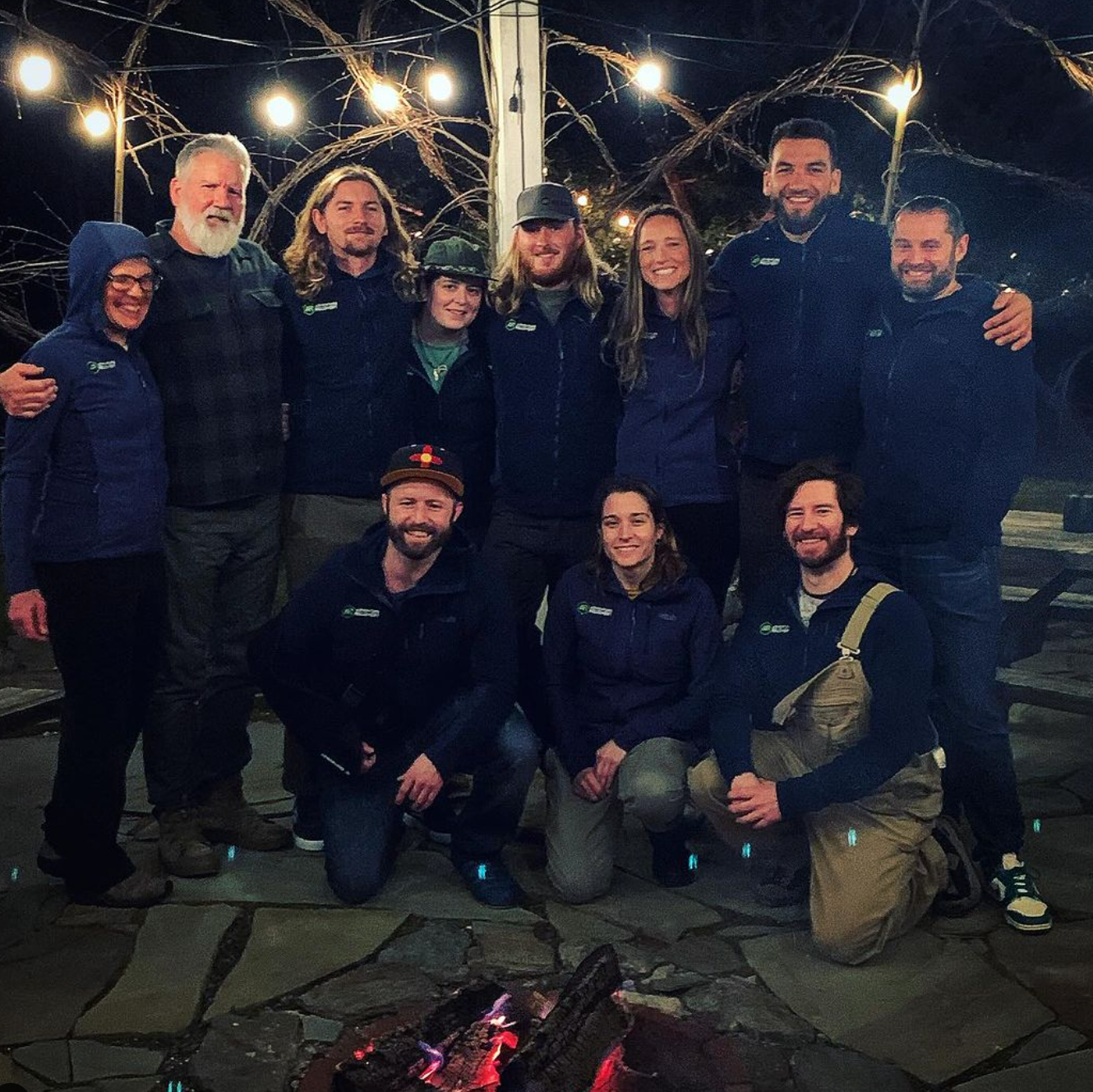 AR Crew holiday party posing together at night around a campfire with string lights overhead, outdoor setting.