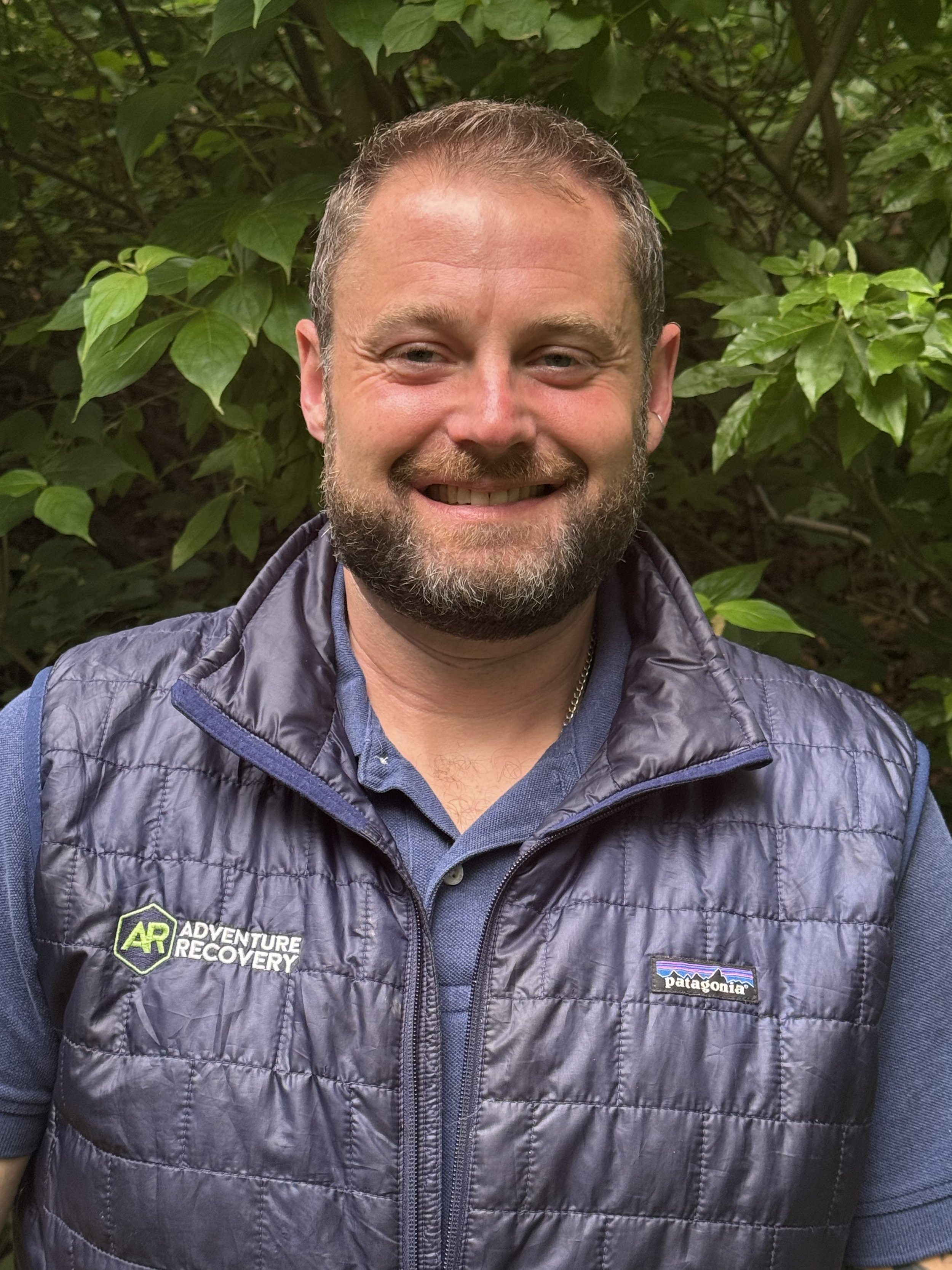 AR executive director, Josh Flaherty, LCSW, standing outdoors in front of green foliage, wearing a navy blue Patagonia vest.