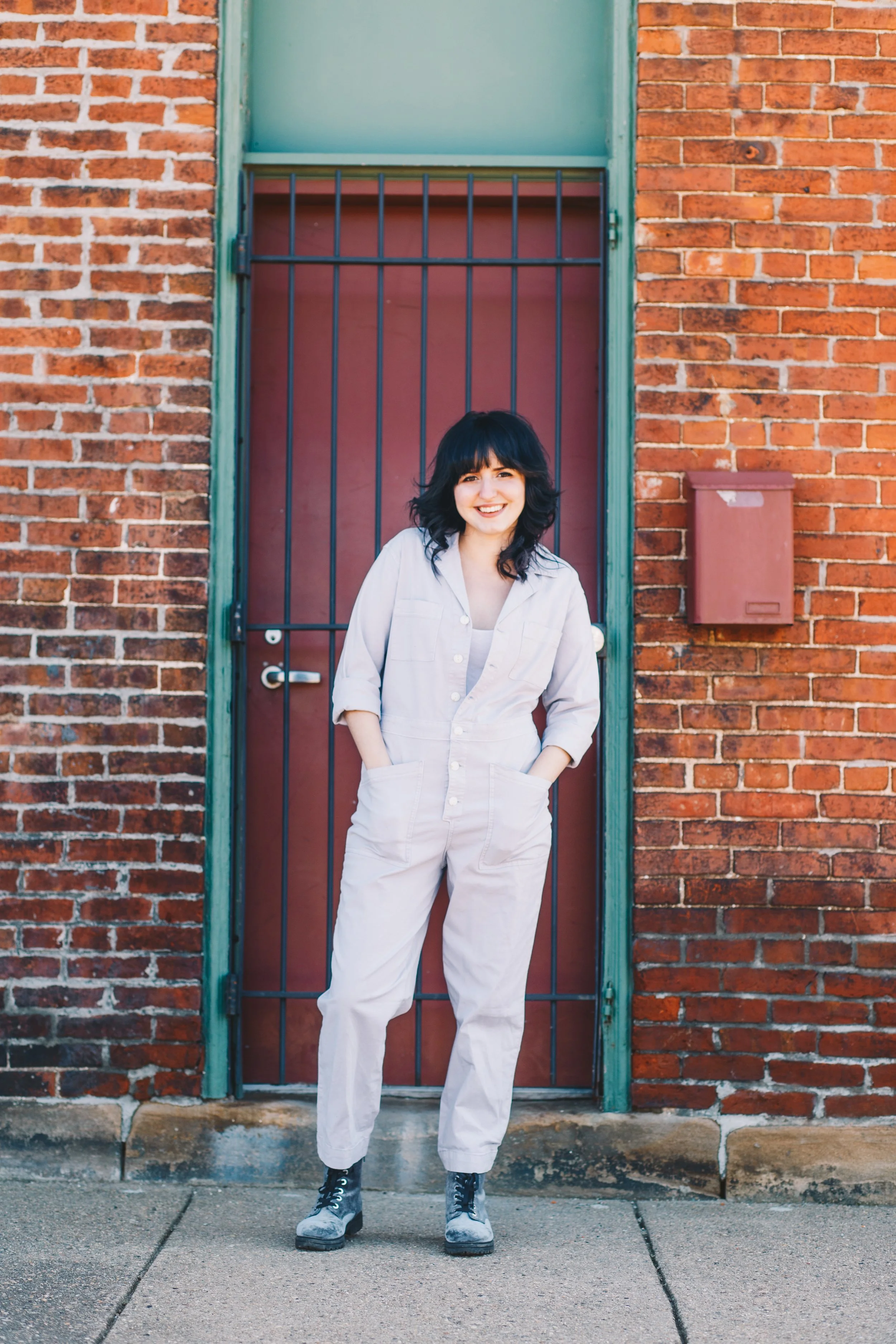 A woman with black hair and bangs smiling and standing with her hands in her pockets in front of a red brick wall and metal barred door, wearing a beige outfit and black boots.