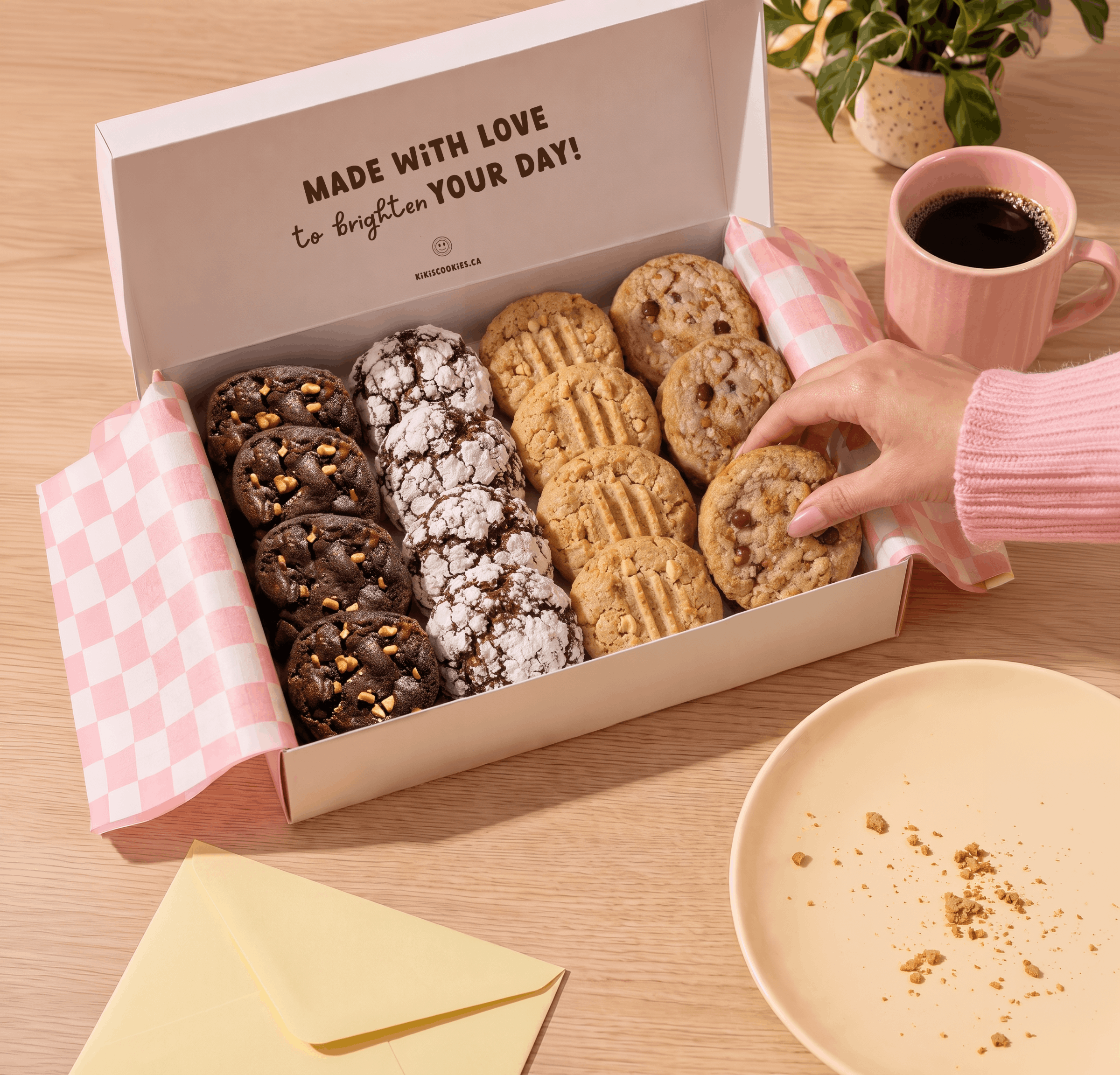 Open Kiki’s Cookies gift box filled with assorted gourmet cookies, with a hand reaching for a cookie beside a pink coffee mug and yellow envelope.