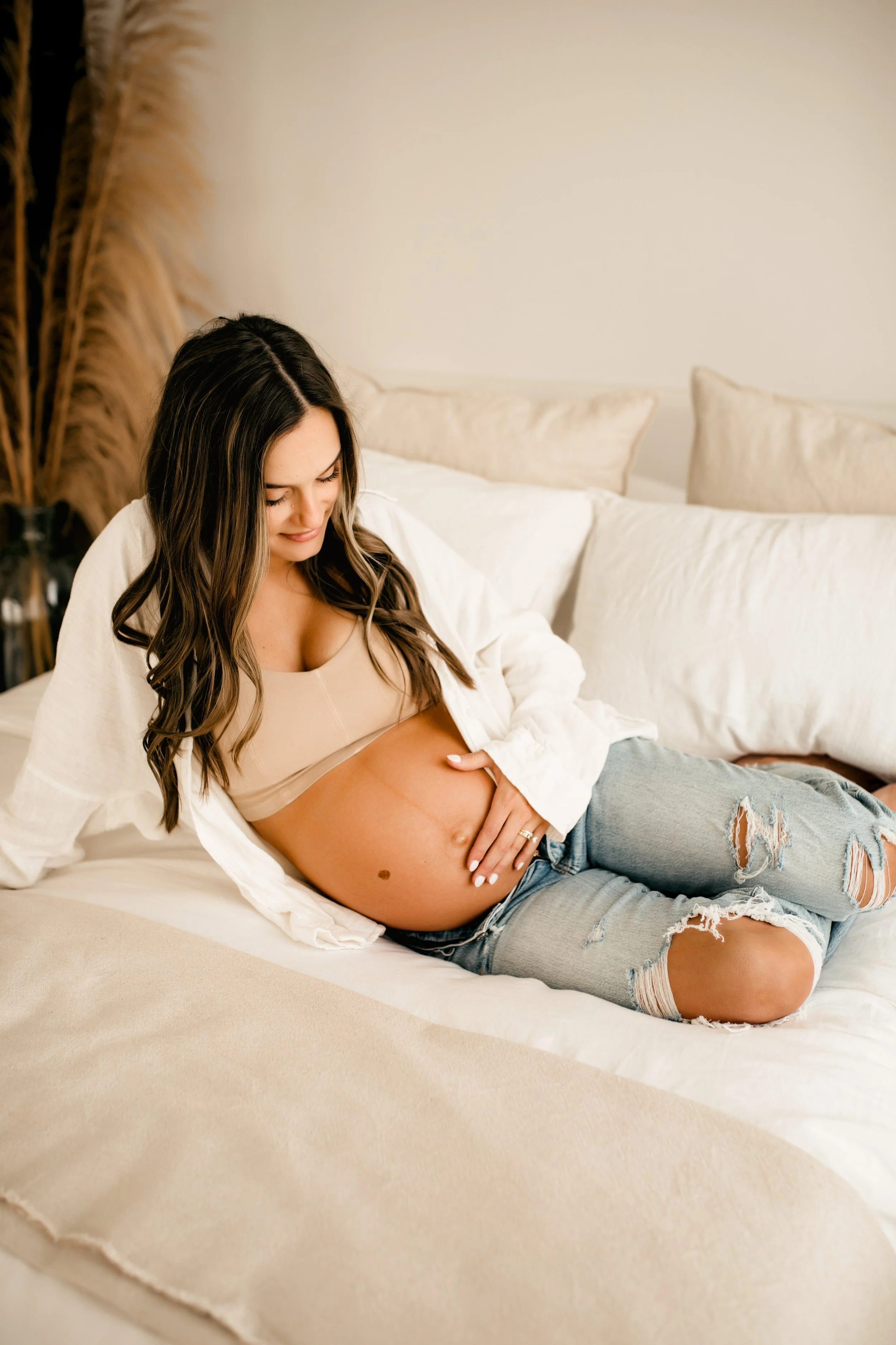 Pregnant woman in jeans resting on a bed, gently holding her belly, with pampas grass decor in the background for her studio maternity session with Jamie Hunt Photography, Nashville's Best Newborn and Family Photographer.