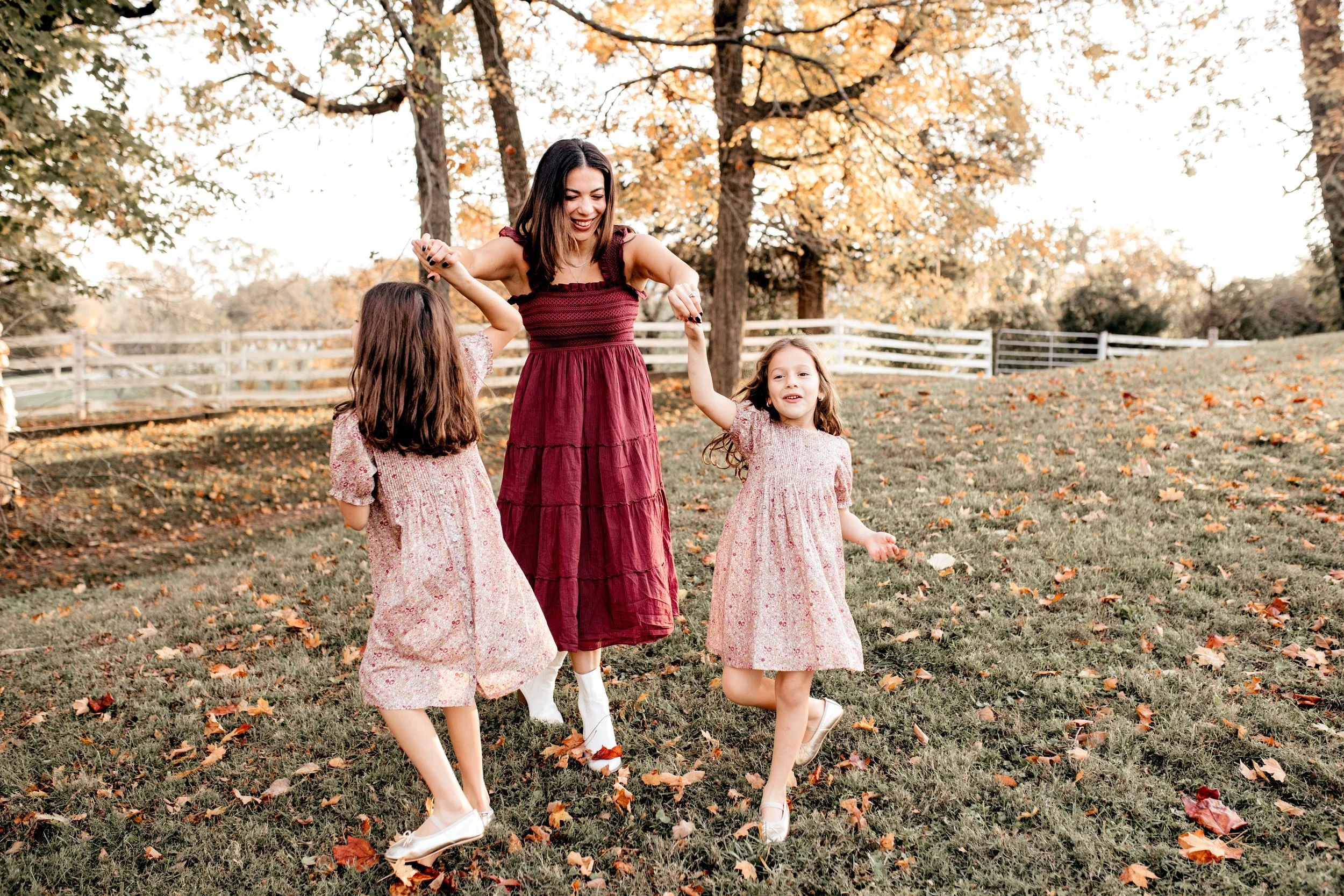 A woman and two young girls playing and holding hands in a park during fall, with trees and fallen leaves around them.   Lifestyle outdoor family session in Nashville, TN