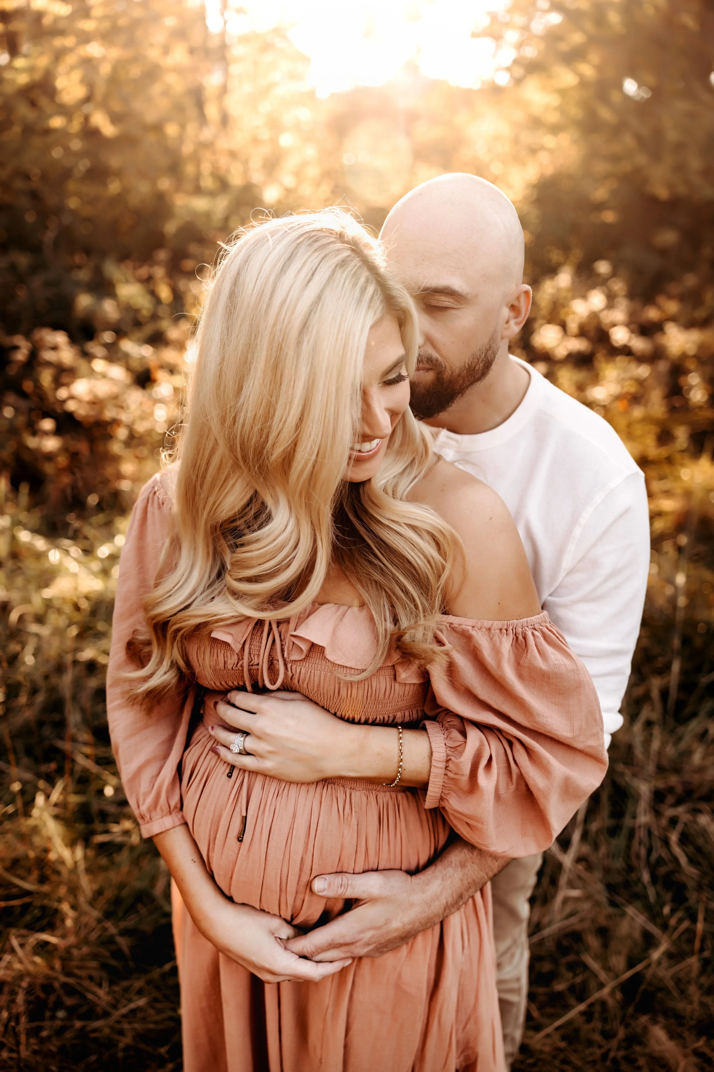 Pregnant couple embracing in a sunny outdoor setting, woman wearing a peach dress for her outdoor maternity session with Jamie Hunt Photography, Nashville's Best Newborn and Family Photographer.