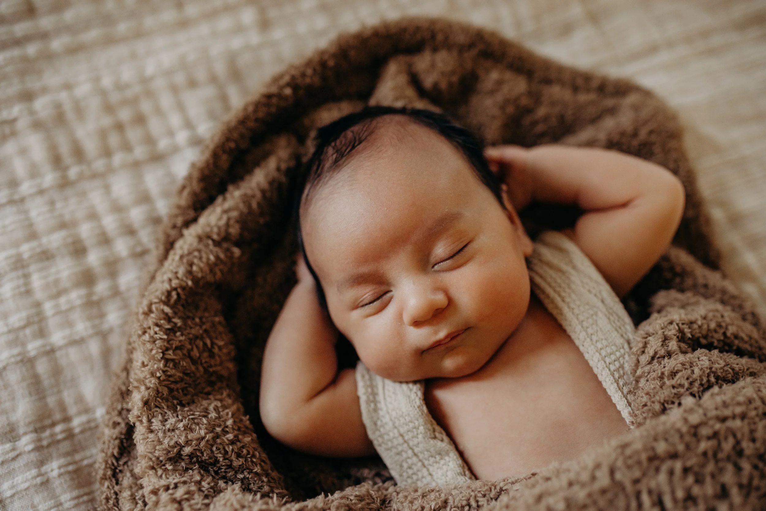 A sleeping baby with short black hair, lying on a soft brown blanket, resting on a beige textured surface.  In-home family newborn session in Nashville, TN