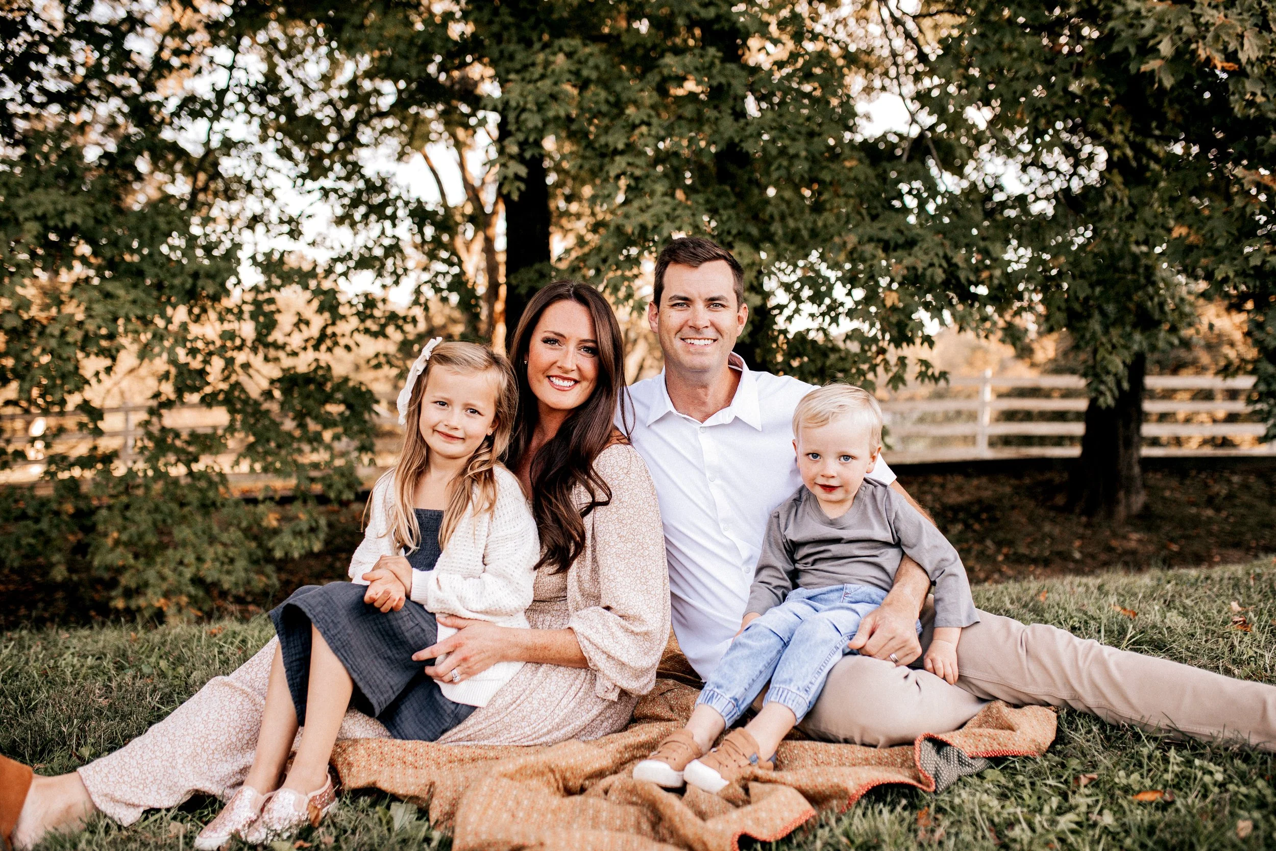 Family of four sitting on a blanket outdoors during sunset, smiling at the camera. The family includes a mother, father, daughter, and son, with a background of trees and a white fence.   Lifestyle outdoor family session in Nashville, TN