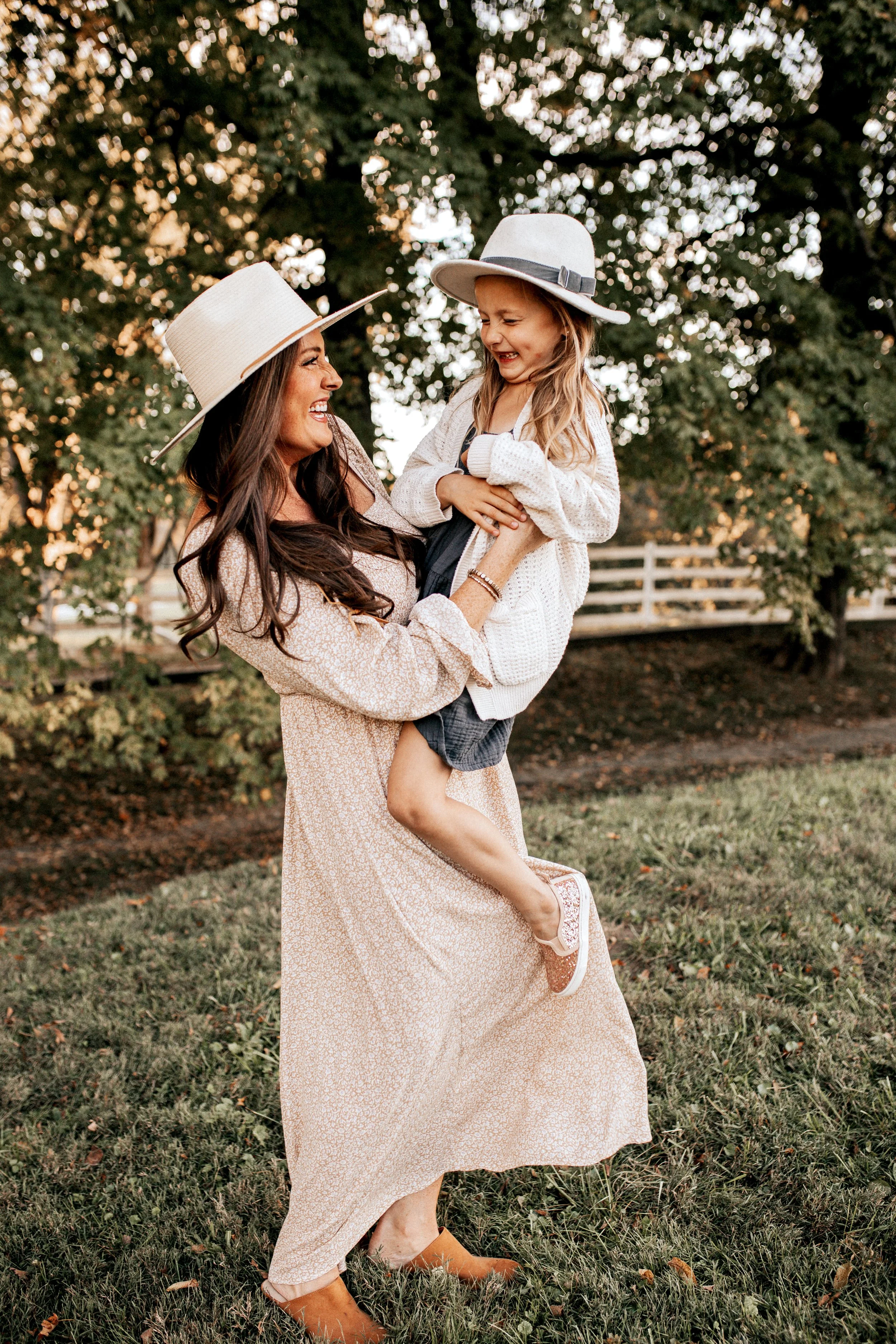 A woman and a young girl wearing wide-brimmed hats outdoors, smiling and laughing while the woman lifts the girl in her arms.  Lifestyle outdoor family session in Nashville, TN
