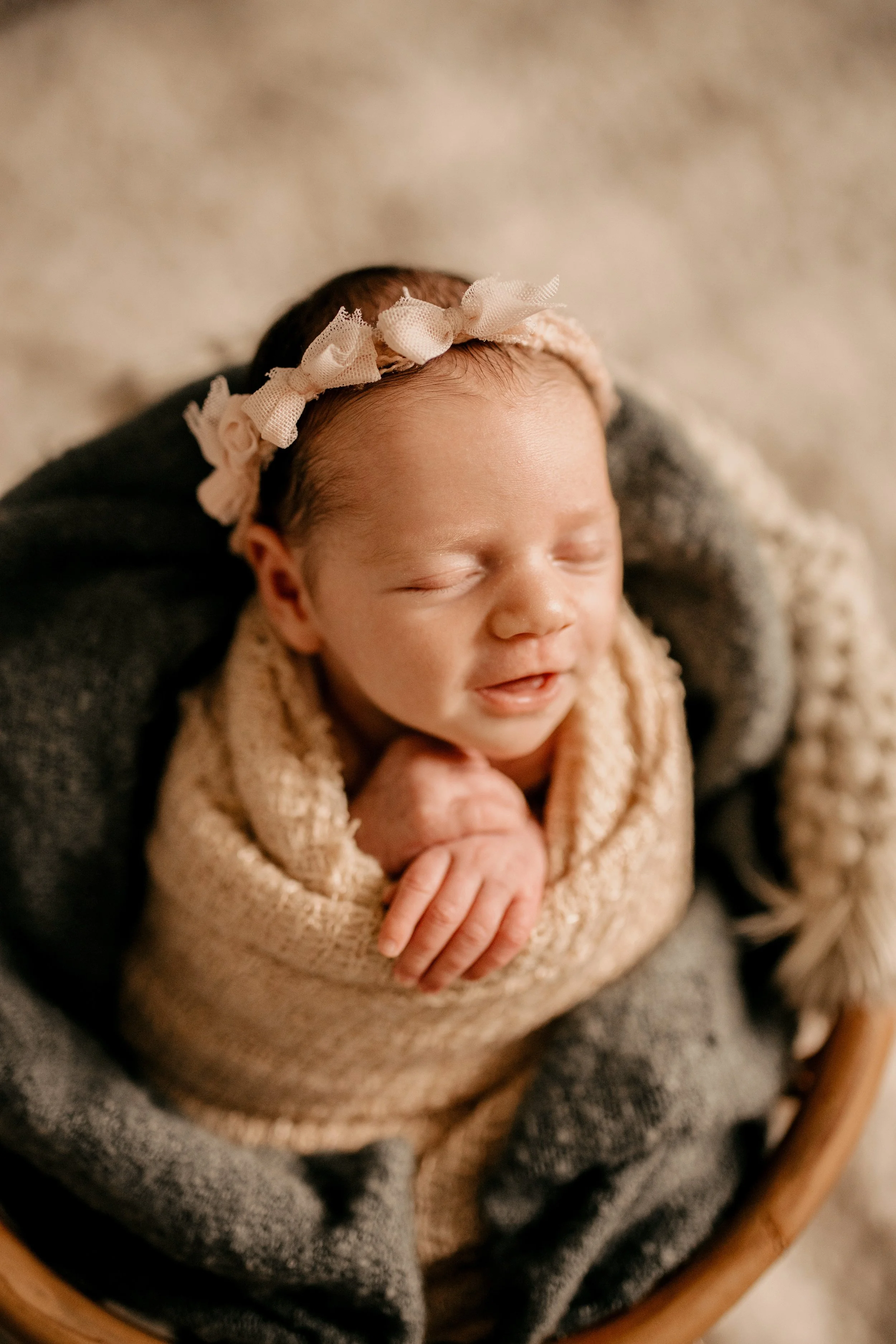 Close-up of a newborn baby girl with a pink flower headband, wrapped in a beige knitted blanket, resting on a gray blanket in a wooden basket.  In-home family newborn session in Nashville, TN