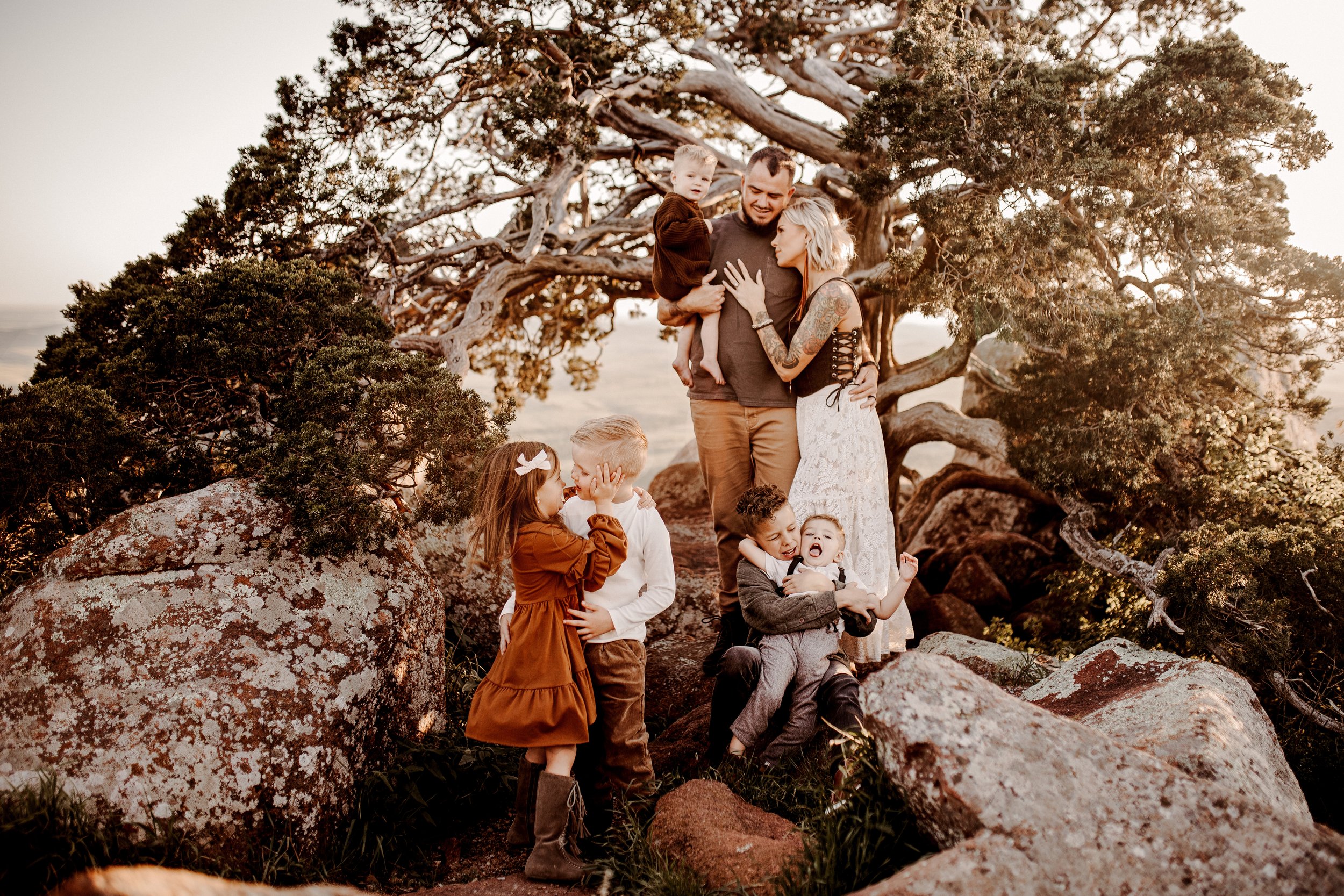 Family of seven on rocks and near trees outdoors at sunset, including two adults and five children, with children interacting and parents holding a child, in a scenic natural setting.  Family Session in Nashville Tn