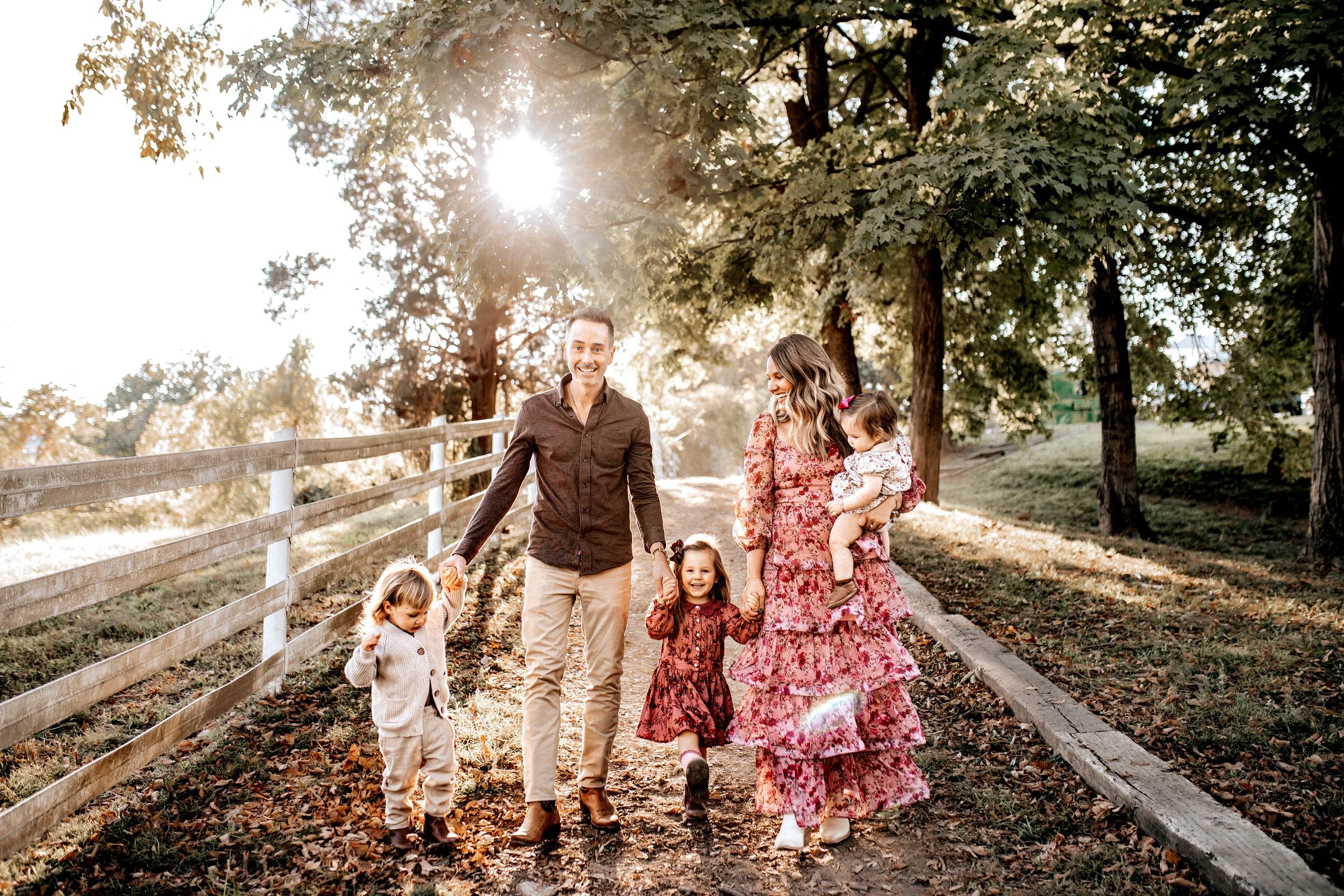 Family of six enjoying a walk on a sunny autumn day along a tree-lined path with fallen leaves, holding hands, smiling and laughing.   Lifestyle outdoor family session in Nashville, TN