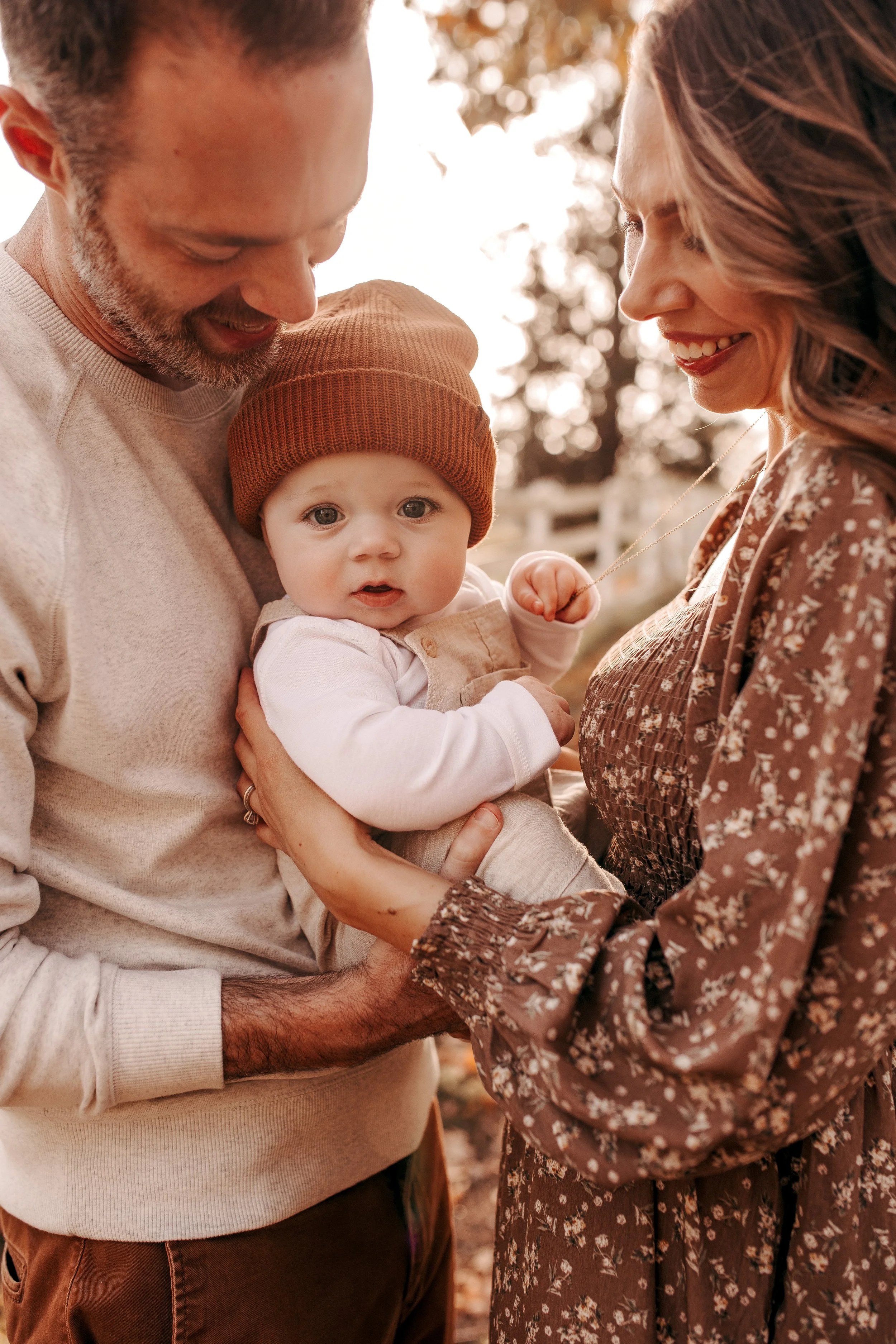 A man and woman holding a baby outdoors in autumn, with trees and a fence in the background, all smiling and dressed in warm clothing.   Lifestyle outdoor family session in Nashville, TN