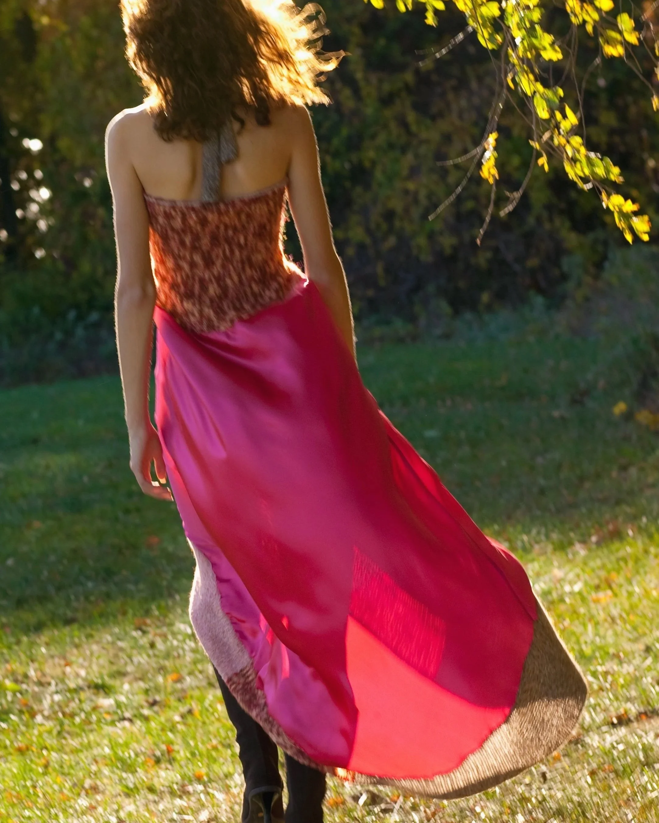 A woman with curly hair wearing a colorful flowy silk dress, walking outdoors during golden hour sunlight, with green trees and grass in the background.