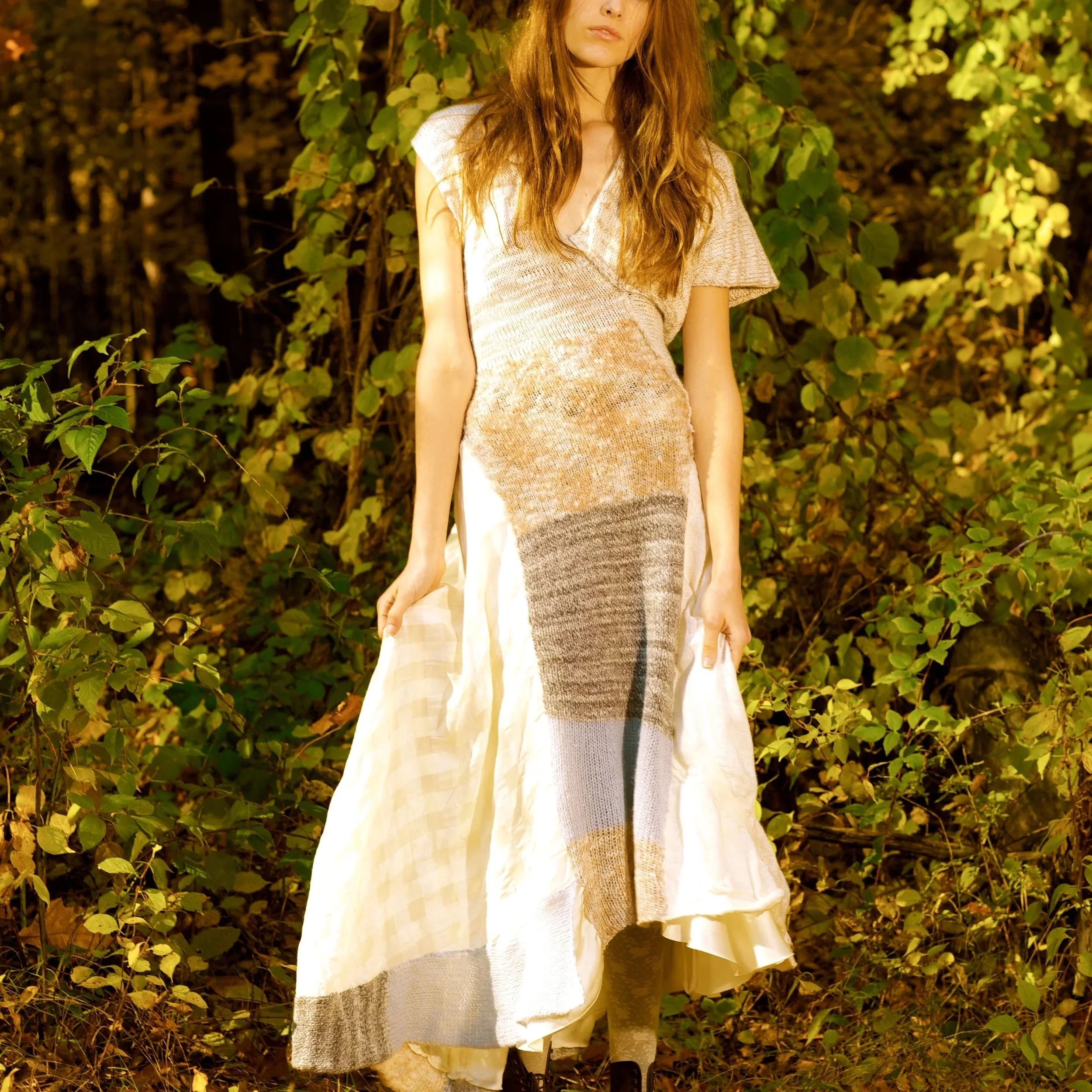 A woman standing outdoors in front of green foliage, wearing a long hand kit dress with a printed pattern resembling abstract art, and holding the sides of her dress.