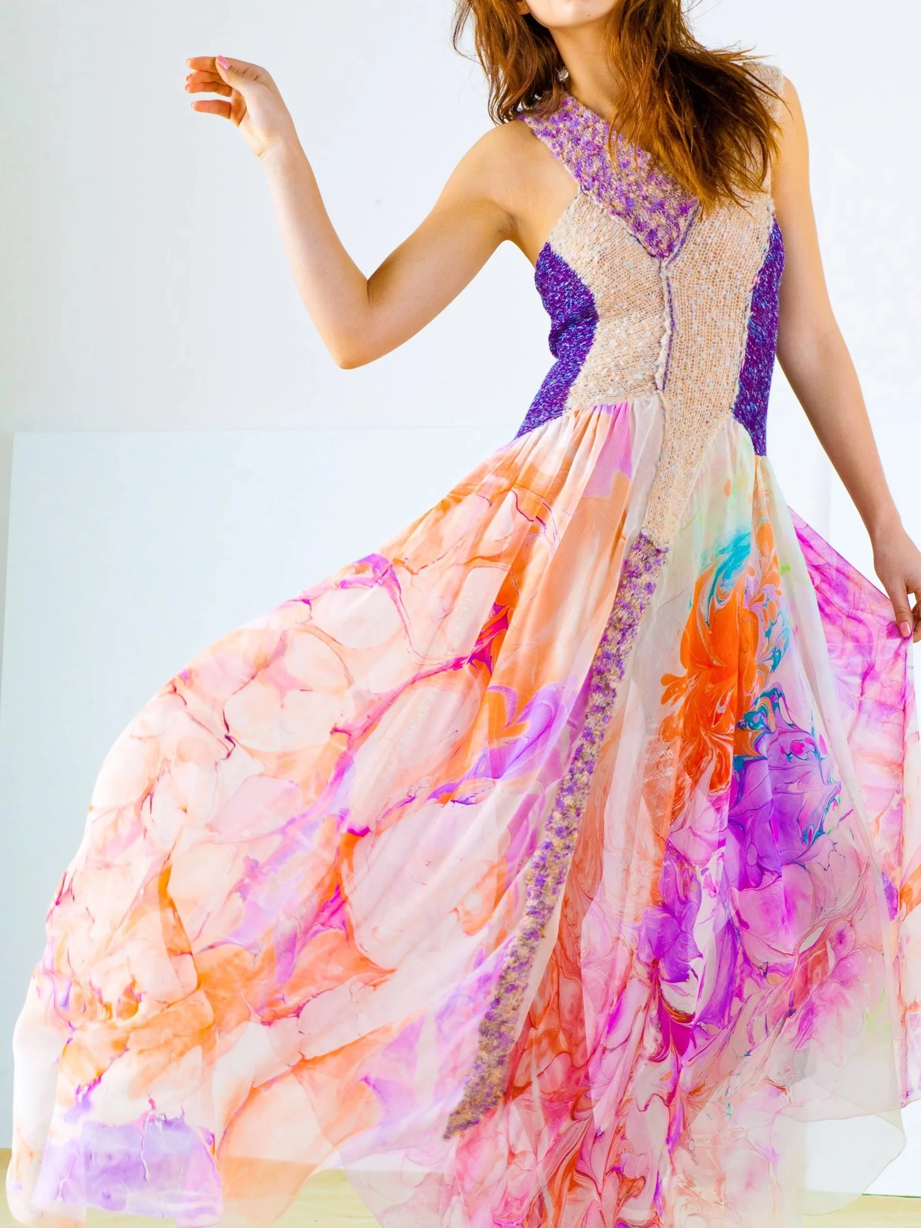 A woman in a colorful, flowing dress with abstract painted patterns and vibrant hues, standing against a plain white background.